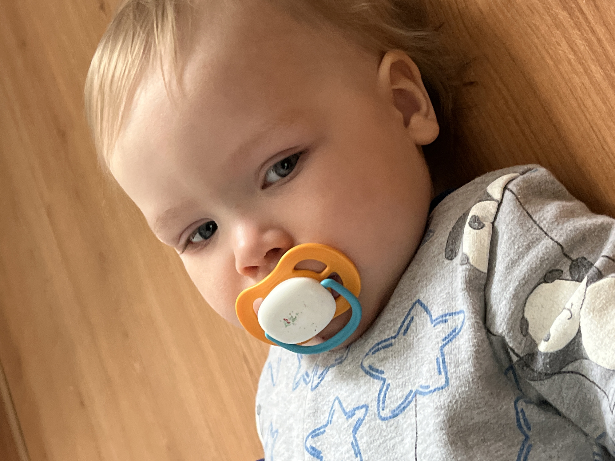 A close-up, slightly angled, high-angle shot shows a baby lying on a light brown wooden surface. The baby is light-skinned with blonde hair and grey-blue eyes. The baby is wearing a grey pajama top with blue star and white panda bear patterns. The baby has an orange and blue pacifier in its mouth. The setting appears to be indoors, and the location is not evident in the image; therefore, it is impossible to verify if it is in Chaikovsky, Russia.