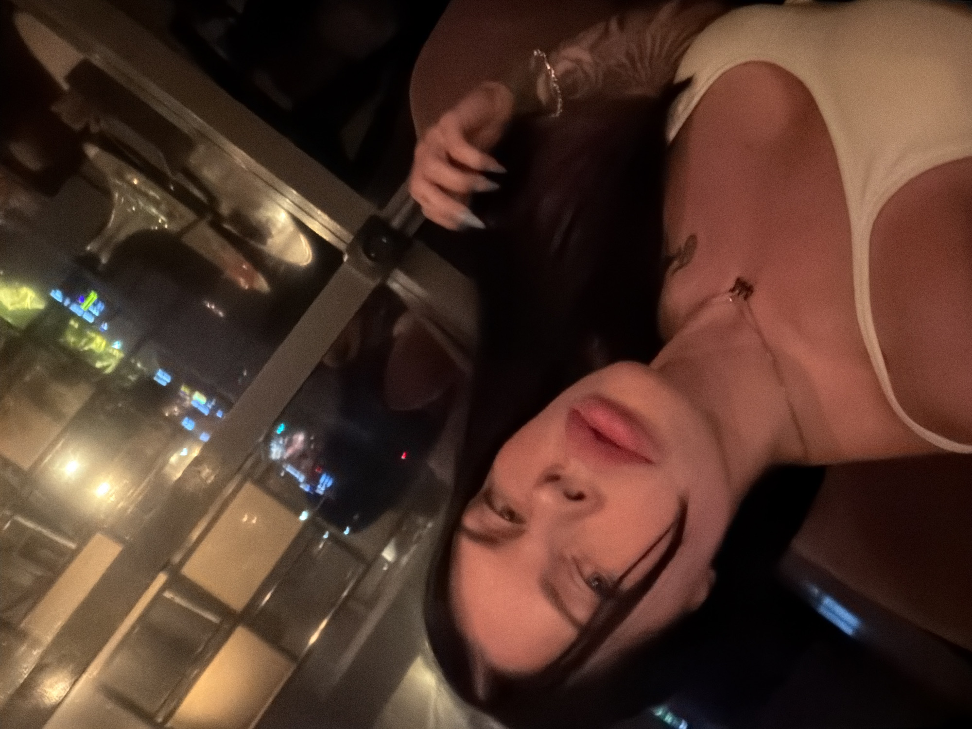 A woman is taking a selfie in what appears to be a dimly lit indoor setting, possibly a restaurant or bar, with reflections of city lights visible through a window. She has dark hair and is wearing a white top. Her lips are full and appear to be glossed. She is wearing a necklace with a small pendant. Her right hand, with light blue-tipped nails, is near her hair. A delicate bracelet is visible on her left wrist, and she has a tattoo on her left forearm. The background shows a blurred view of tables and chairs, and neon lights, indicative of a nighttime urban environment. There is no specific information in the image to suggest the location is Ufa, Russia.