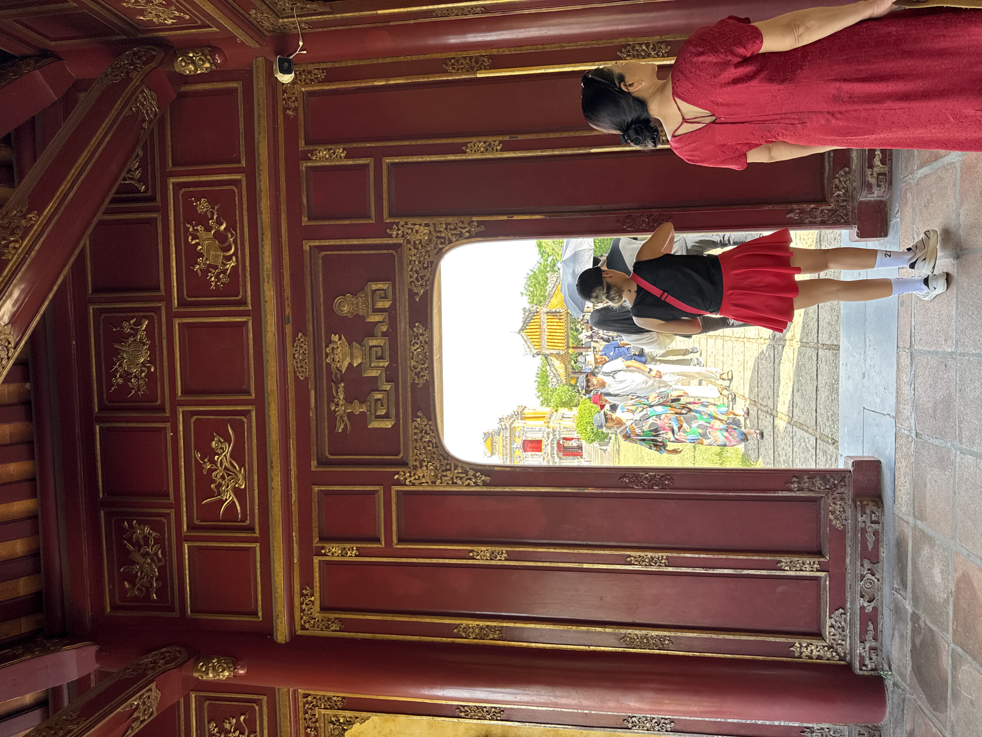 The image depicts a view through a large, ornate red doorway with gold trim and intricate carvings. The doorway leads to an outdoor scene where several people are walking on a paved path in front of traditional Vietnamese architecture. The architecture visible through the doorway features yellow roofs with curved edges and red accents, characteristic of historical buildings in Phú Xuân Ward, Vietnam, likely within the Imperial City of Huế.
In the foreground, standing just inside the doorway, are two women. The woman on the right is wearing a red dress. The woman on the left is facing away from the camera, wearing a black top, a red skirt, and white sneakers with socks. She has a red strap across her shoulder and is looking out at the scene beyond the doorway. Her back is partially obscured by a black umbrella held by someone further inside the doorway.
Beyond the doorway, several individuals are walking towards the camera. One woman in the middle ground wears a colorful patterned dress and a hat. Other figures are visible, including someone in a white shirt and a person with a blue shirt. The ground outside the doorway is paved with stone tiles, and there is green grass and trees in the background. The overall lighting suggests it is a sunny day. A security camera is mounted on the wall to the right of the doorway, overlooking the interior space.