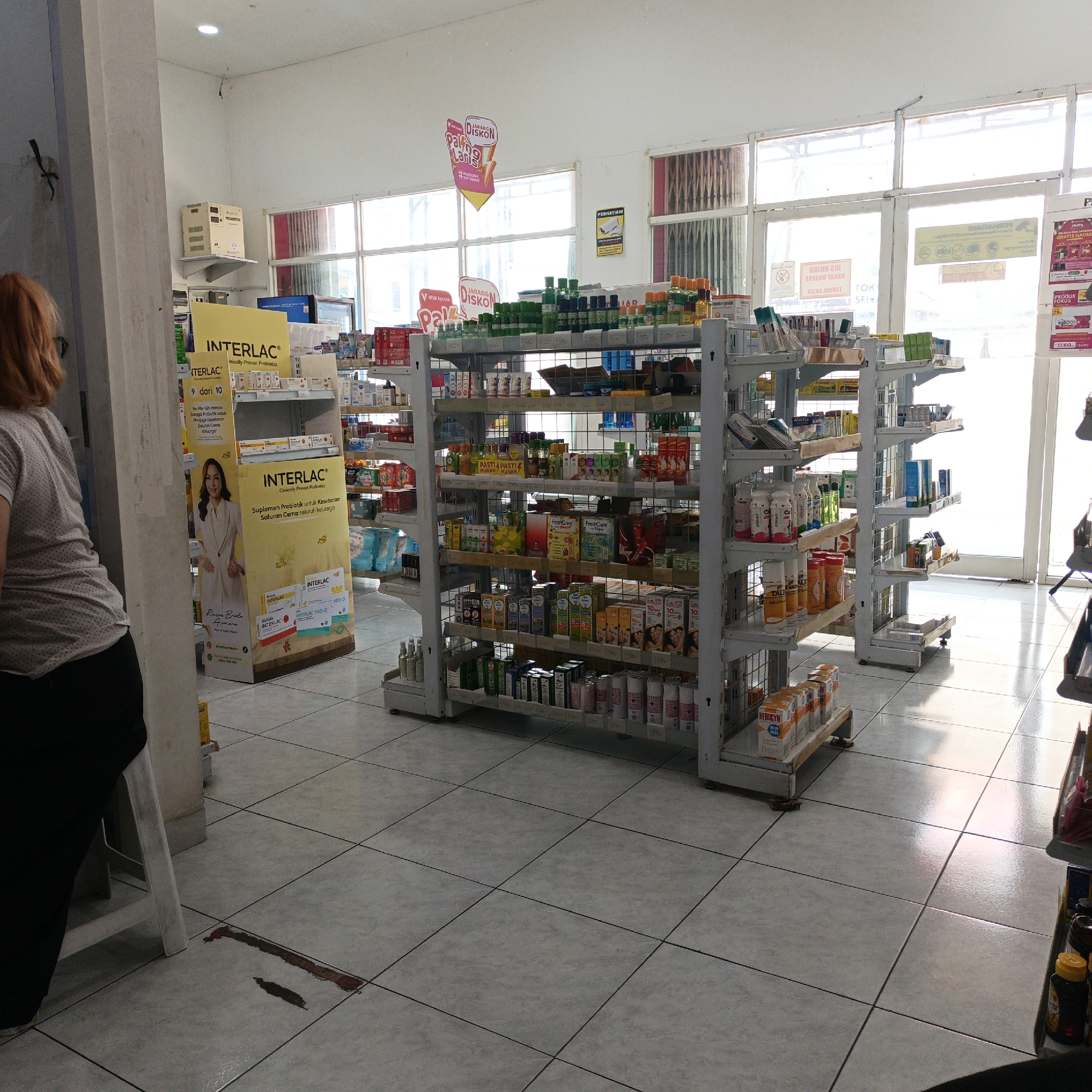 The image depicts the interior of a pharmacy in Semarang, Indonesia. The pharmacy is well-stocked with various health and personal care products displayed on metal shelving units. In the foreground, a woman with short, reddish-brown hair is partially visible from the back, wearing a light-colored, short-sleeved, polka-dot shirt and black pants. She appears to be standing near a pillar.
The shelves are organized with products such as bottles of medicine, vitamins, supplements, and other pharmaceutical items. Several promotional posters and displays are visible, including a large yellow display for "INTERLAC® Clinically Proven Probiotics" which features an image of a woman. The floor is tiled with a grey, square tile pattern. Light streams in from large windows located at the back of the pharmacy, creating bright reflections on the floor. In the far background, a closed metal grate or shutter is visible. There is a spill of reddish-brown liquid on the floor near the pillar in the foreground, and some water spillage near it.