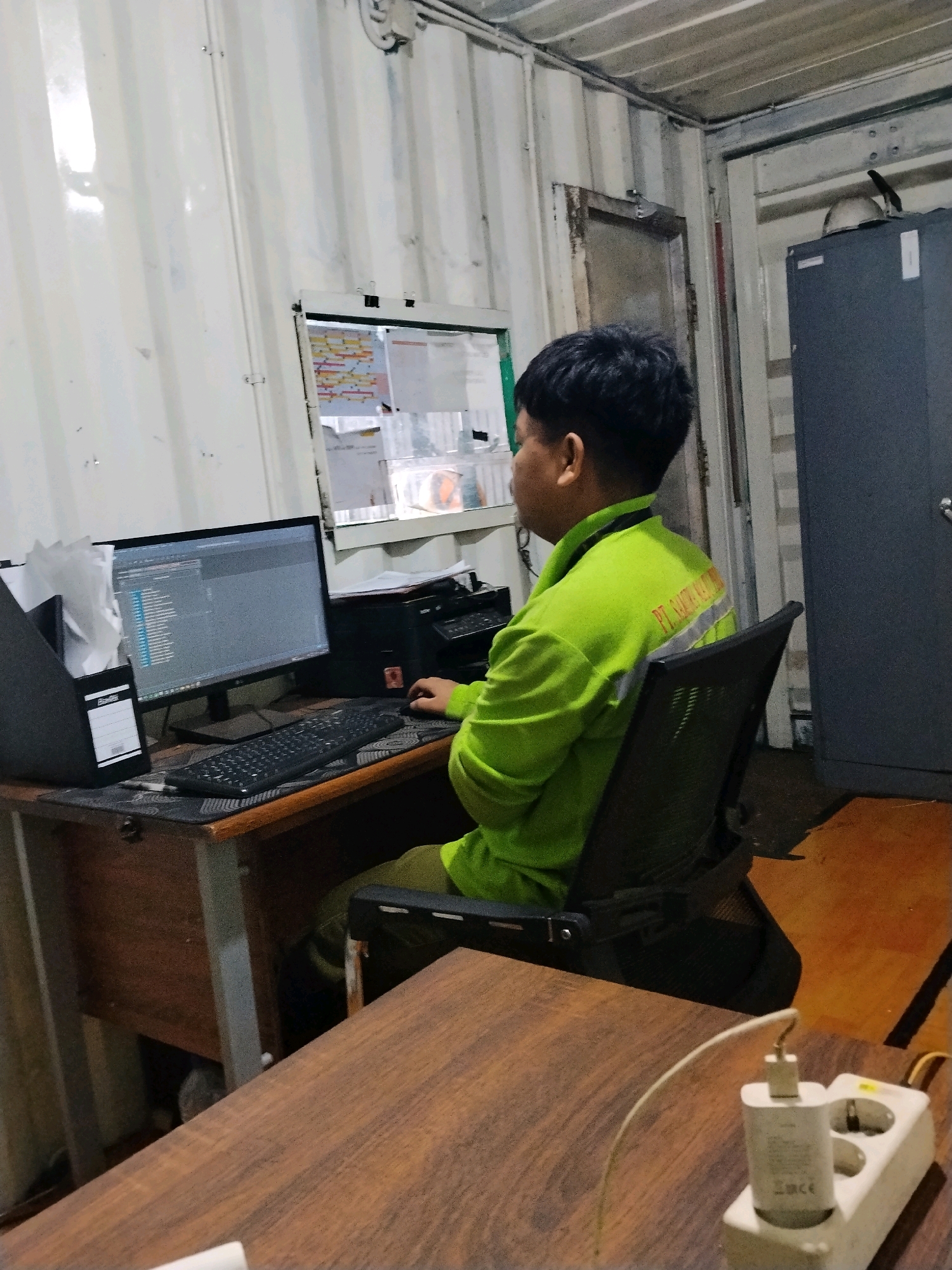 A man is sitting at a desk in what appears to be a shipping container converted into an office, in Sempayau, Indonesia. He is wearing a bright green reflective jacket with text on the back that reads "PT. SAHAT KAHURIPAN". The man is looking at a computer monitor and has his right hand on a mouse. On the desk are a keyboard, a printer, a black folder with papers, and a white tissue dispenser. The office has corrugated metal walls and ceiling. A large dark gray cabinet is visible on the right side of the room. In the foreground, a wooden table has a white electrical adapter plugged into a socket, with a white cable attached.