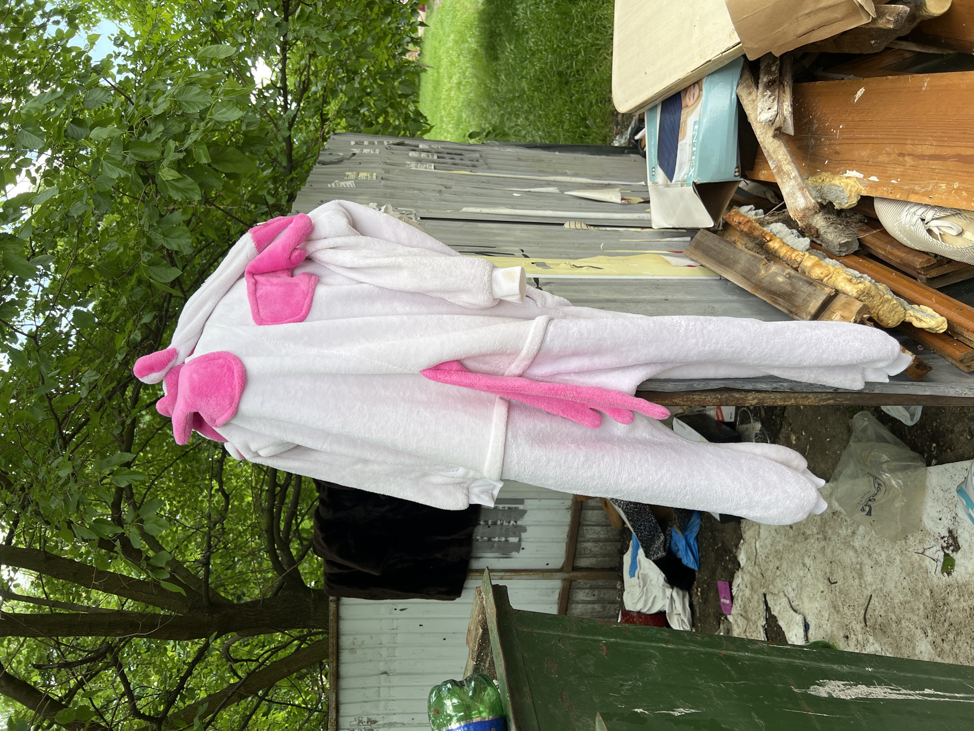 The image depicts a white and pink unicorn onesie lying on top of a cluttered metal structure. The structure appears to be part of a dilapidated outdoor area with a green bin and various discarded items. The background includes trees and a glimpse of a grassy lawn. The exact location cannot be ascertained to be Krasnodar, Russia, as there is no information in the image to suggest this.