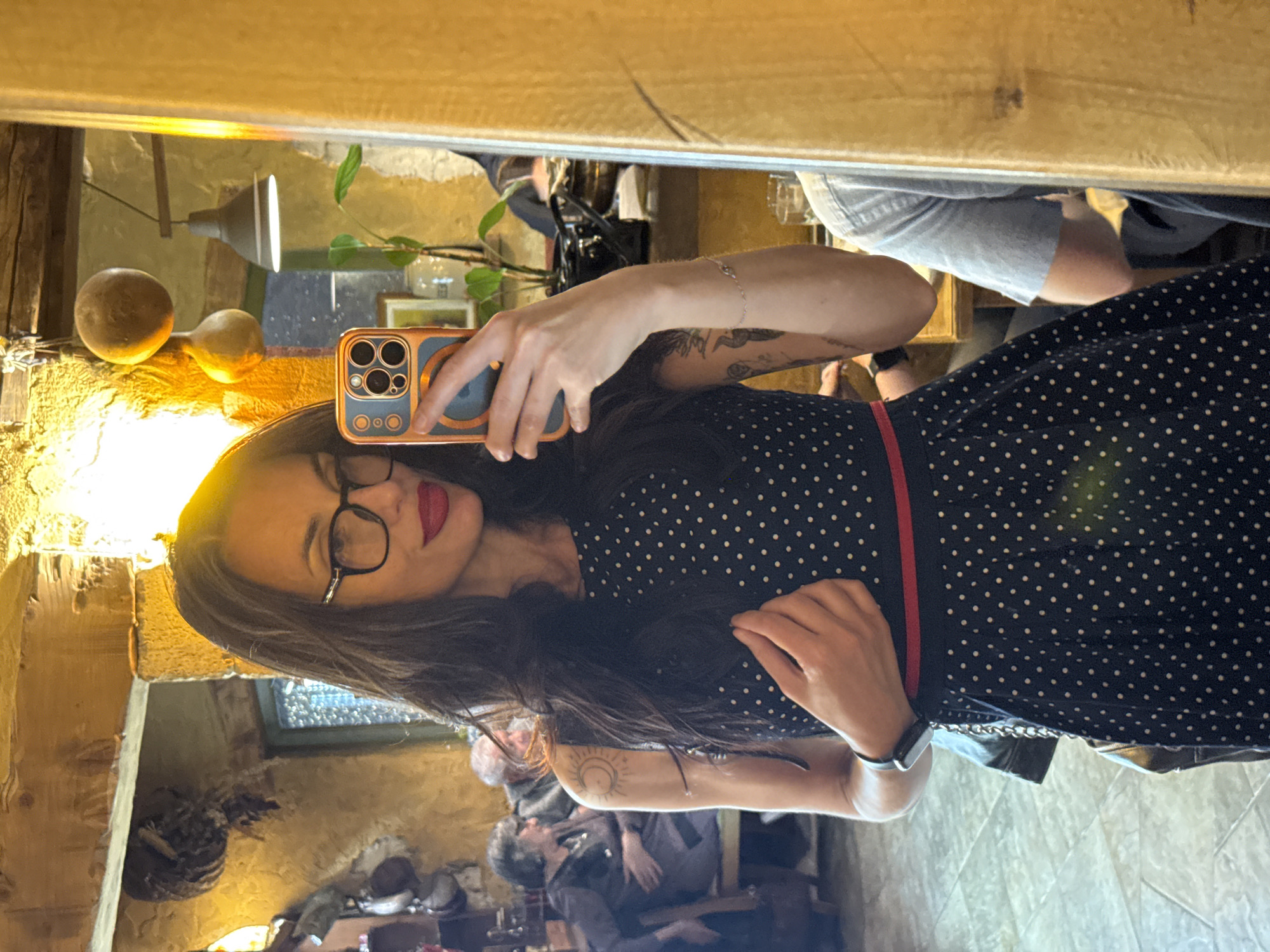 The image shows a woman in a black and white polka dot dress taking a selfie in a mirror. She is wearing glasses and has dark hair. In the reflection, other people can be seen seated. The environment appears to be indoors, possibly in a restaurant or café. The background features rustic-style decor with wooden beams and textured walls. The image may have been taken in Pleven, Bulgaria.