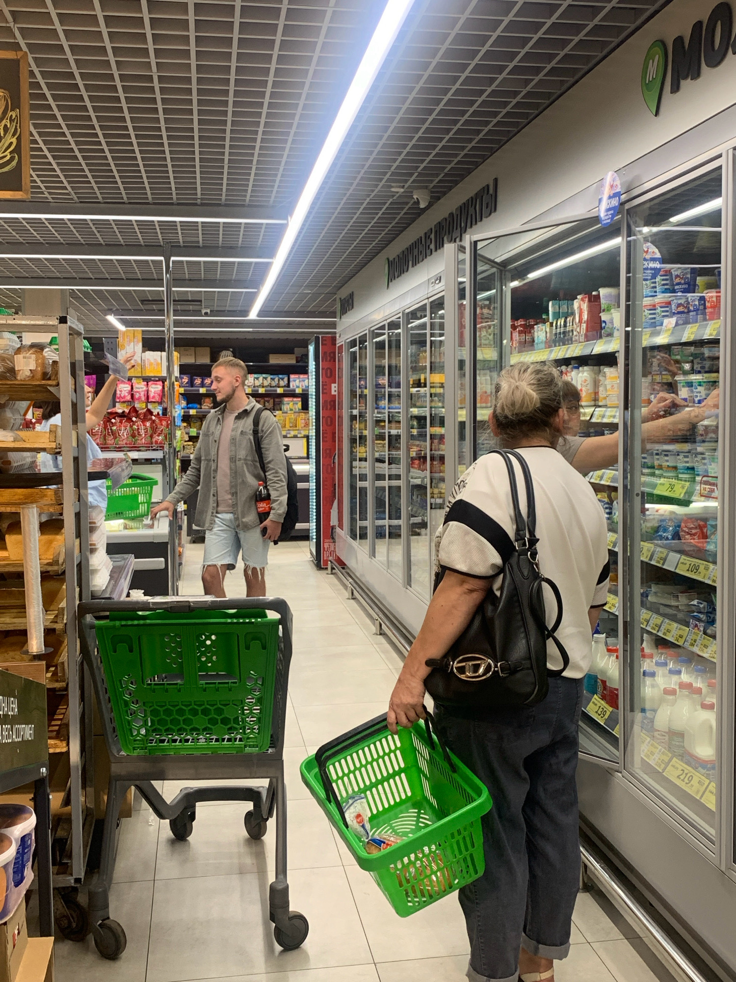The image captures an interior view of a brightly lit supermarket aisle. In the foreground, a person is walking away from the camera, holding a green plastic shopping basket in their right hand. They are wearing dark pants and a white top with black trim. To the left, a large green shopping cart is partially visible.
In the middle ground, a man with short blonde hair is standing in the aisle, wearing a grey jacket over a tan t-shirt and light blue denim shorts. He is holding a bottle of cola in his right hand and a phone in his left. He appears to be interacting with someone to his left, who is partially obscured by shelving.
Further down the aisle, a row of refrigerated display cases filled with dairy products lines the right side of the image. The wall above the display cases features signage in Russian, including a logo with the letter "M" and the word "МОЛОЧНЫЕ ПРОДУКТЫ" (Dairy Products). The ceiling is a grid of metal panels with integrated fluorescent lighting. The floor is tiled.
The scene is consistent with a modern grocery store environment in Russia, such as a "Magnit" supermarket, which is a common chain in cities like Prokopyevsk. The overall atmosphere is one of everyday shopping activity.