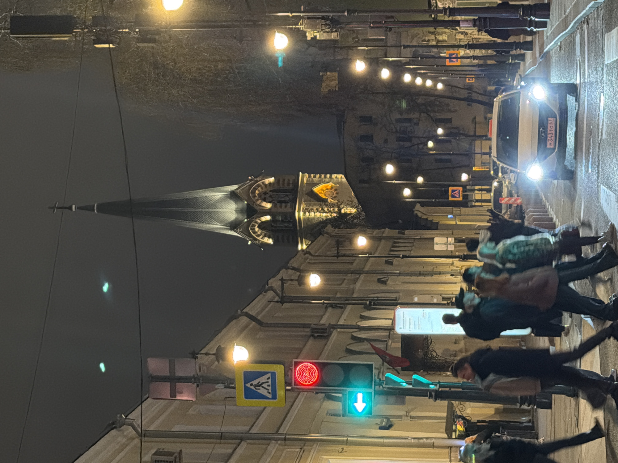At night in Moscow, Russia, people cross a street at a green light near a traffic signal with a pedestrian crossing sign. A tall building with a spire is visible in the background. A white vehicle with the license plate M343TO67 is traveling down the street. Streetlights line the street.