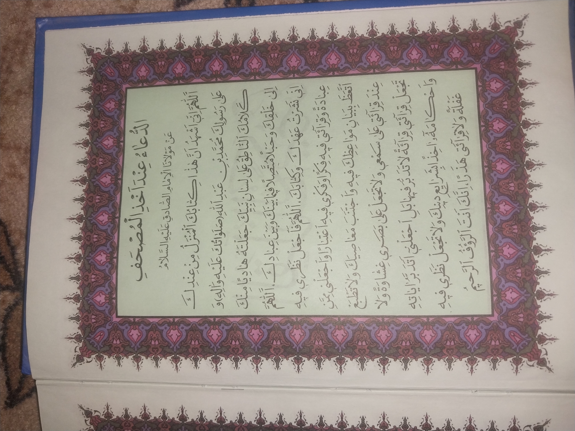 The image shows an open book with Arabic text, framed by ornate floral borders in shades of purple, pink, and dark red. The text appears to be religious in nature, possibly a prayer or supplication. The book is placed on a textured surface, which is brown and appears to be fabric, possibly a rug or blanket. The lighting suggests an indoor setting, with the light source coming from the left. There are no people or animals visible in the image. The location of Blagoveshenka, Kyrgyzstan, is not depicted or suggested by any visual elements in the image.