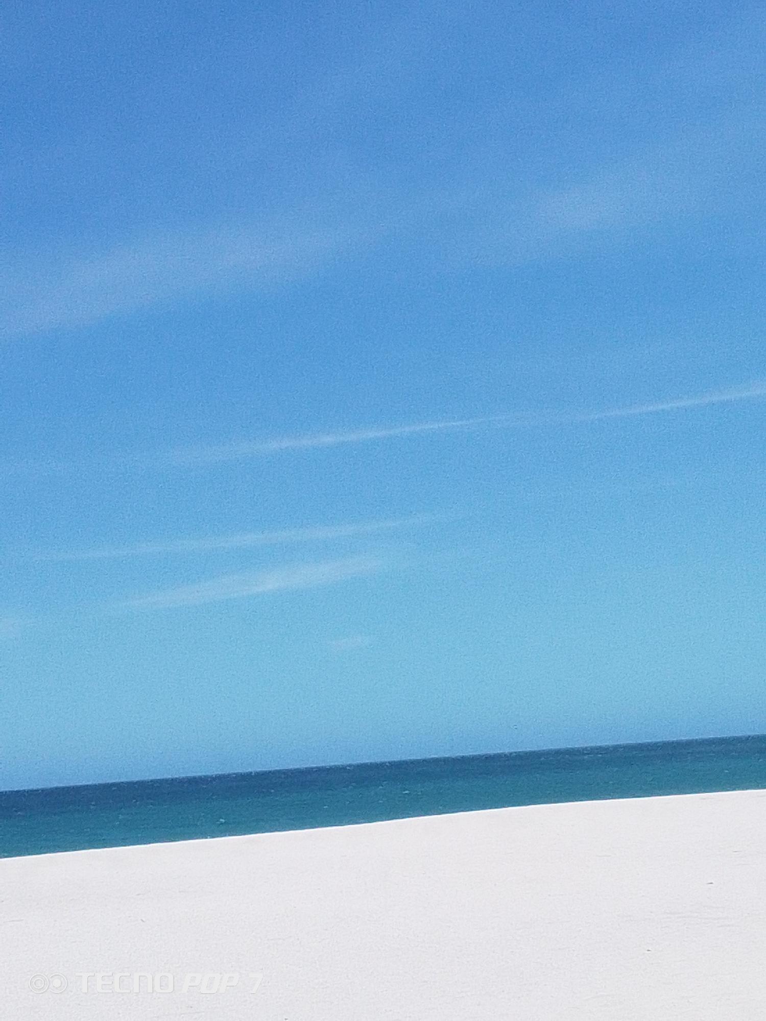 The image depicts a serene coastal scene, likely taken in Pedro González, Venezuela. A clear, bright blue sky dominates the upper portion of the frame, with subtle wisps of clouds visible. Below the sky is a strip of dark blue ocean water, and in the foreground, a white sandy beach extends into the distance. There are no people or objects visible in the scene beyond the natural elements. The horizon line between the ocean and the sky is distinct. The image presents a minimalist composition, emphasizing the natural beauty of the coastal environment.