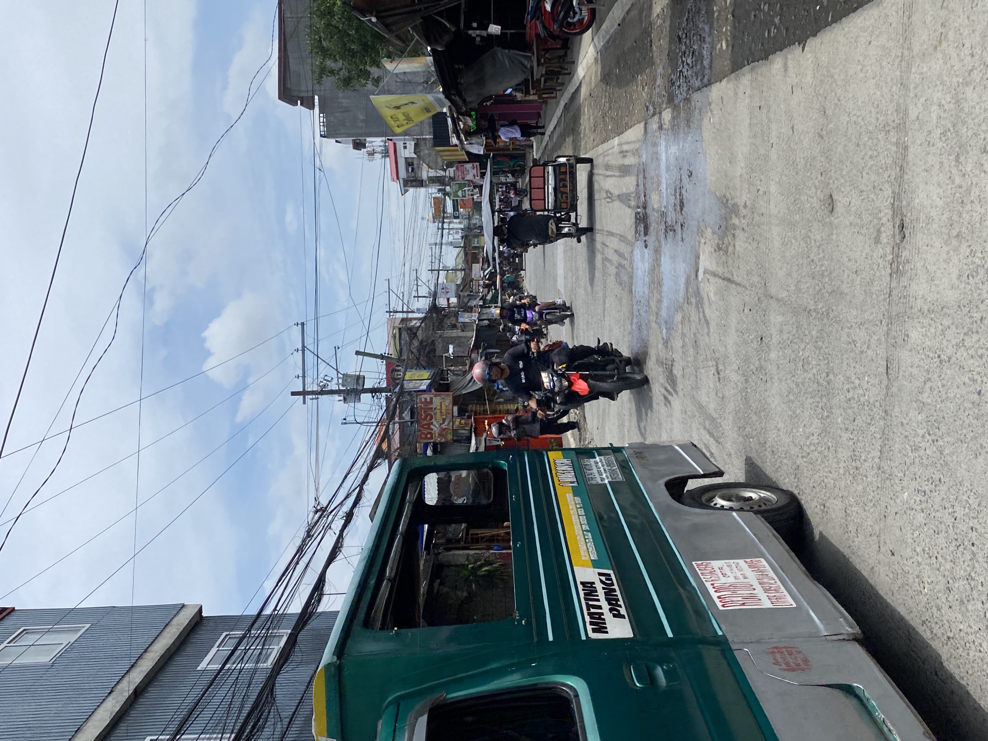 The image depicts a street scene in Davao City, Philippines. In the foreground on the left, a green and silver van with "MATINA PANGI" written on its side is partially visible. The van appears to be a passenger vehicle, possibly a jeepney or similar public transport. The street is paved with concrete and shows signs of wear, with some wet patches and tire marks.
Several people are present, primarily on motorcycles and a tricycle. On the right side of the street, a man wearing a helmet and a dark shirt is riding a red motorcycle. Further down the street, other motorcycles and a tricycle are visible, some carrying passengers. The buildings lining the street appear to be a mix of commercial establishments and residential structures, with some shops open and displaying goods. Electrical wires are strung overhead, a common sight in urban areas of the Philippines. The sky is partly cloudy, indicating daylight conditions. The overall impression is a busy, everyday scene in a Philippine city.