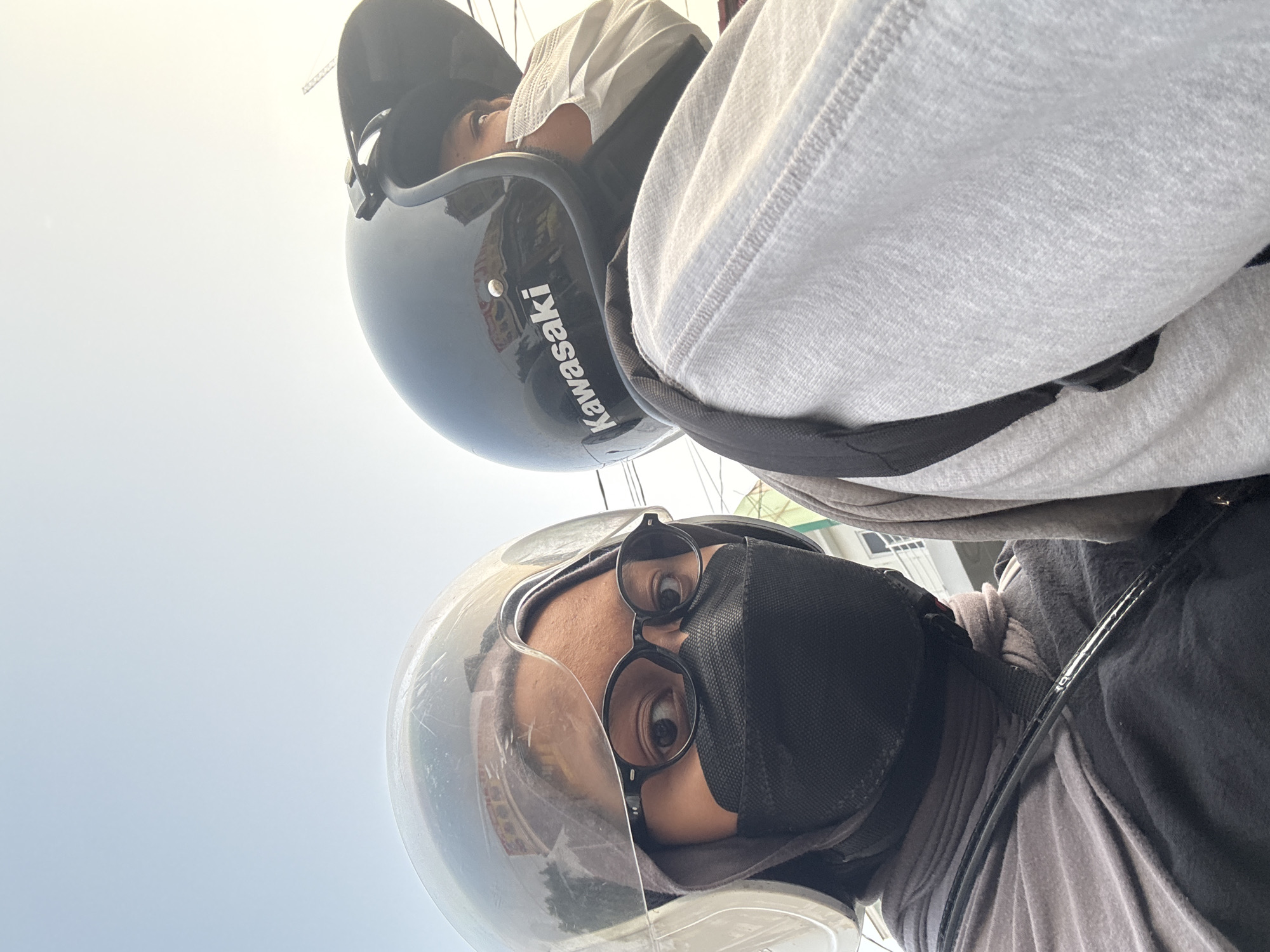 The image shows a close-up, upward-angle selfie of two individuals wearing helmets and face masks, likely on a motorcycle in West Jakarta, Indonesia. The person in the foreground, on the left, wears a transparent bubble helmet, round glasses, a black mask, and a gray headscarf and outer garment. Their eyes are visible, looking directly at the camera. The person behind, on the right, wears a black helmet with the "Kawasaki" logo visible and a white disposable mask. They are wearing a light gray hoodie. The background shows a bright sky with some faint reflections of buildings visible in the foreground helmet's visor. The image captures a moment of travel, emphasizing personal protective gear for riding.