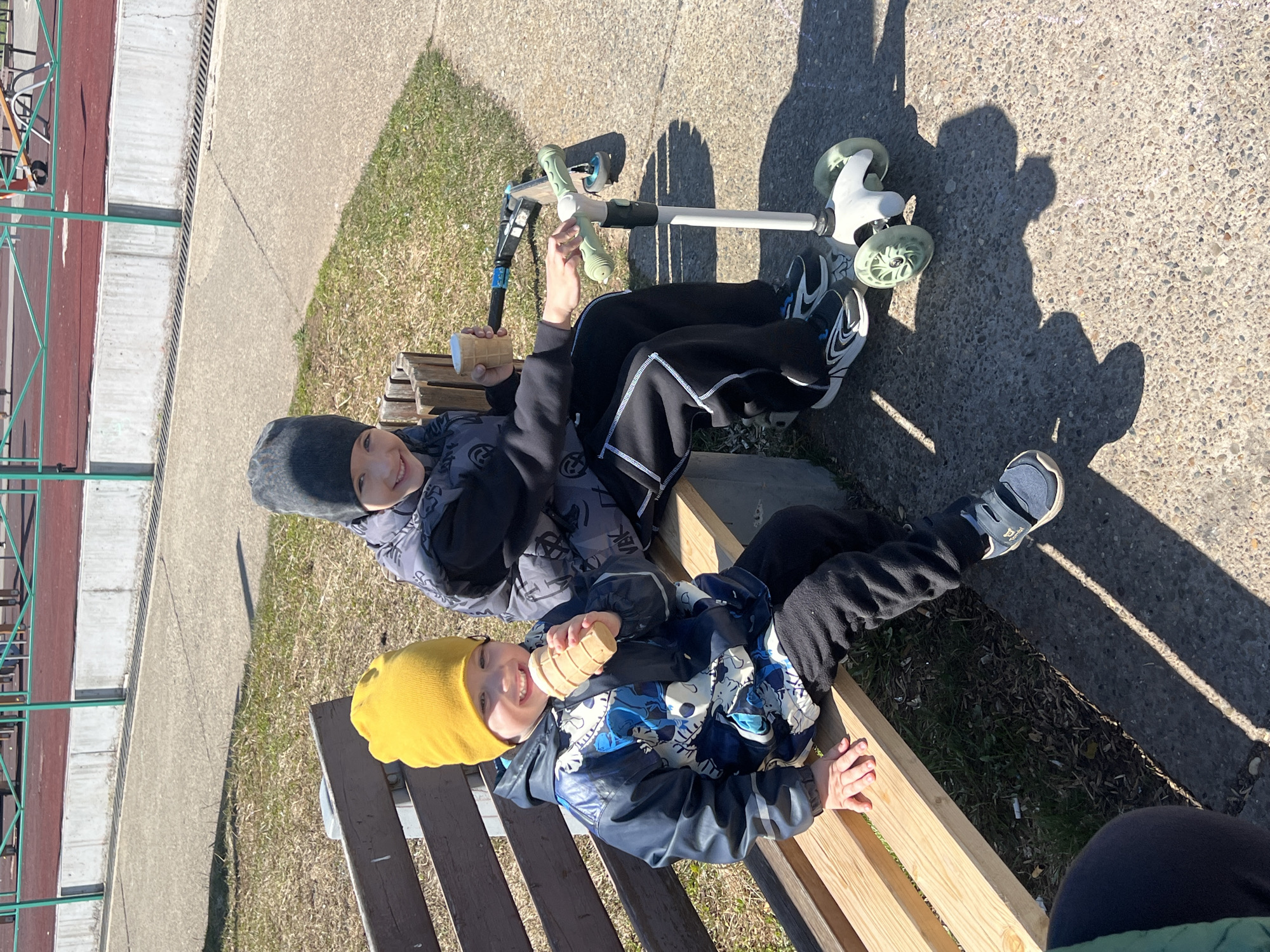 Two boys are sitting on a wooden bench in what appears to be a park. The boy on the left wears a gray beanie and a jacket with a patterned design, holding a cylindrical object. The boy on the right wears a yellow beanie and a blue jacket, also holding a cylindrical object. They are positioned near two scooters on a concrete surface. Grass and other park elements are visible in the background. The location is in Марковский, Russia.