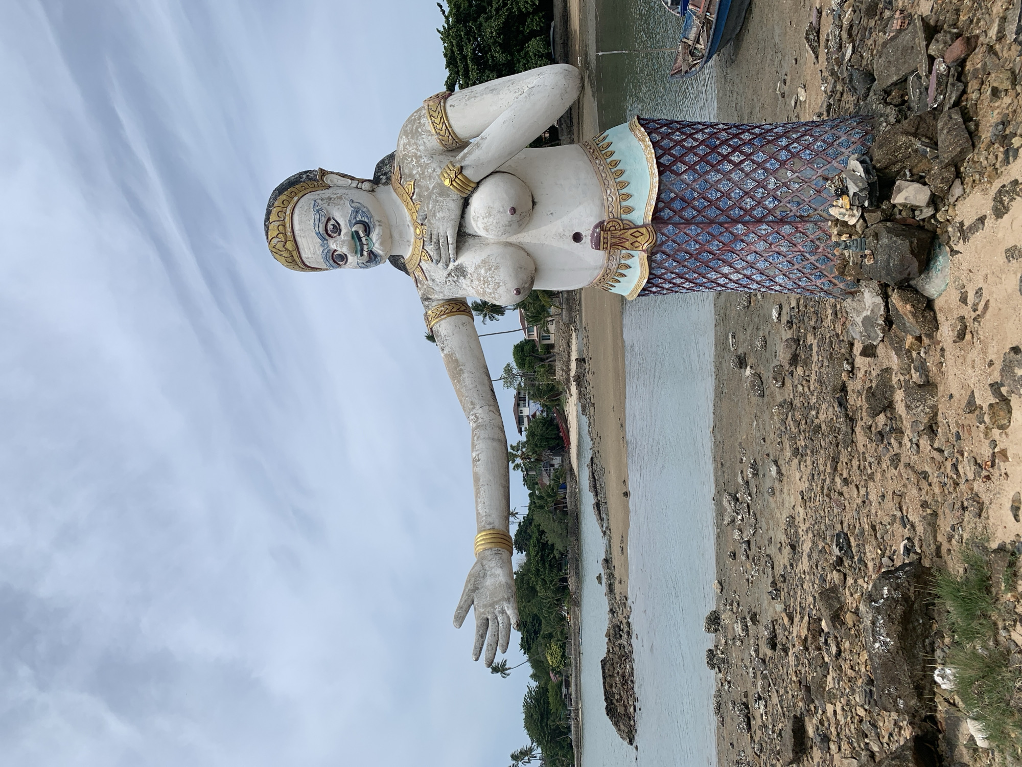 A full-body statue of a mythical creature, possibly a Yak, stands on a rocky, sandy beach in Ko Samui District, Thailand. The statue is white with gold and blue accents, and wears a gold headdress and bracelets. Its lower body is covered in a blue, patterned skirt that resembles scales, suggesting it may be a mermaid or Naga. The statue is positioned facing left, with its left arm extended outwards as if in welcome. The background features a body of water, a sandy shoreline with scattered rocks, palm trees, and some buildings. The sky is overcast with light gray clouds. A small blue boat is visible to the right of the statue. The statue is set on a base decorated with offerings of small figurines and rocks.