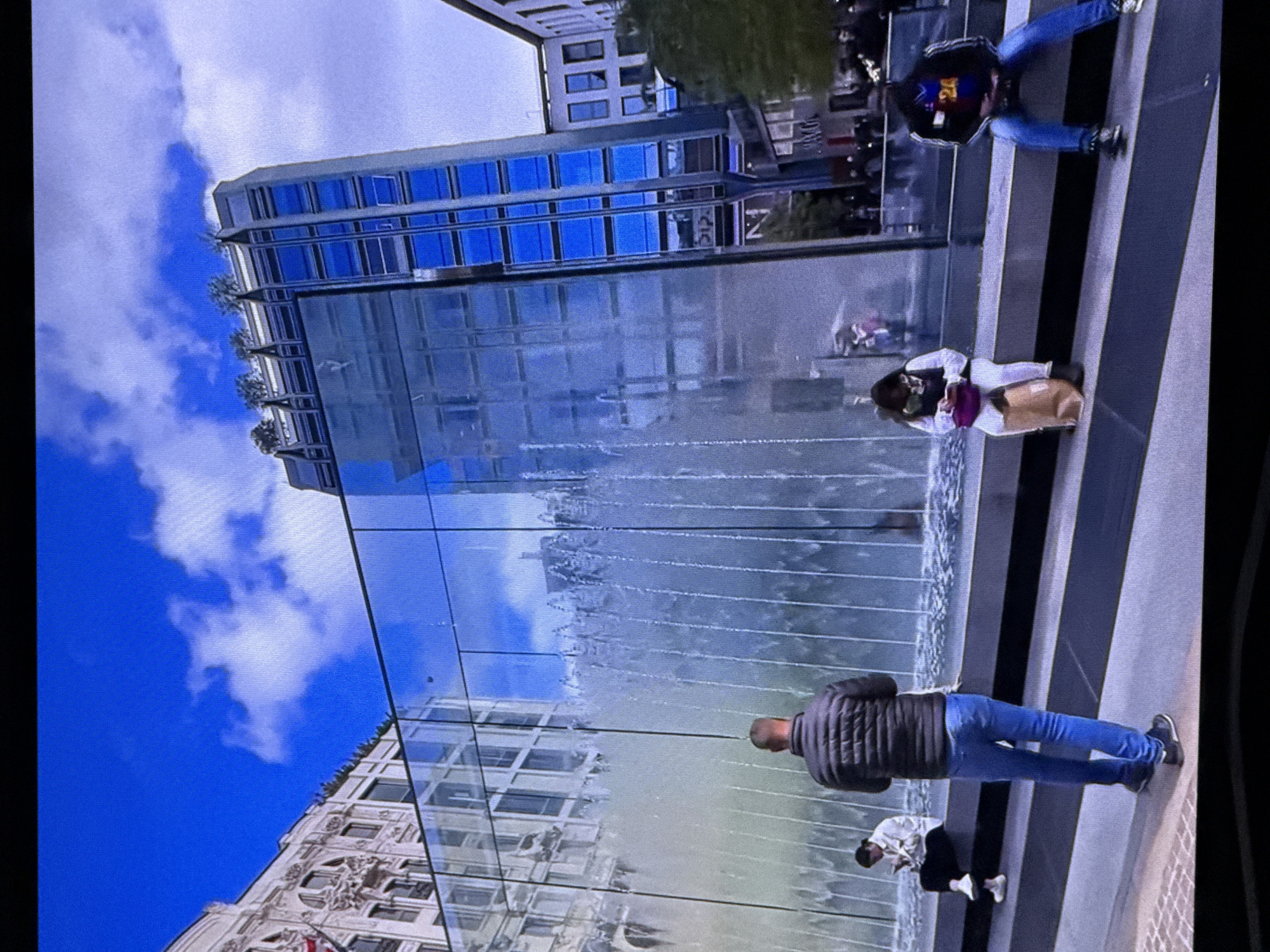In Dubai, United Arab Emirates, a street scene features a modern glass building with a cascading waterfall facade. Several individuals are present in the foreground. A man in a black puffer jacket and blue jeans stands facing away from the camera, near the waterfall. To his left, a person in a white top and dark pants sits on a ledge, looking at their phone. Further to the right, another person sits on a ledge wearing a jacket with the number 24 on it, blue jeans, and sneakers. The background shows more buildings and a few trees. The sky is blue with scattered clouds.