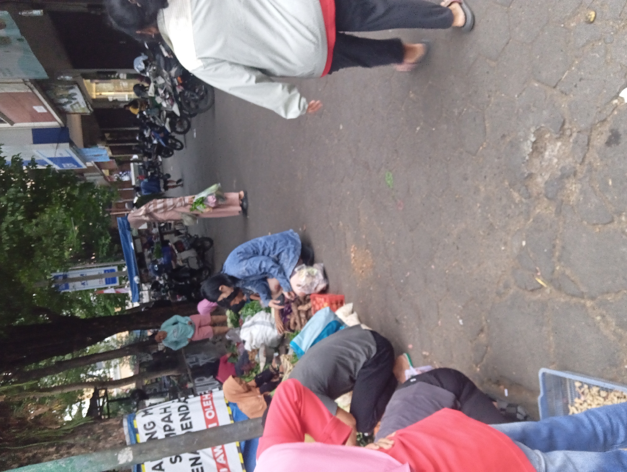 The image captures a street scene in Yogyakarta, Indonesia, likely a bustling market area given the presence of vendors and customers. In the foreground, several individuals are engaged in what appears to be a transaction or preparation of goods, with some crouching and others standing. A person in a red shirt is prominently visible on the left, with another in a gray shirt next to them. In the background, more people are present, along with parked motorcycles, indicating a common mode of transport in the region. Buildings line the street, suggesting an urban setting. The overall atmosphere appears dynamic, with a mix of commercial activity and daily life unfolding on the pavement.