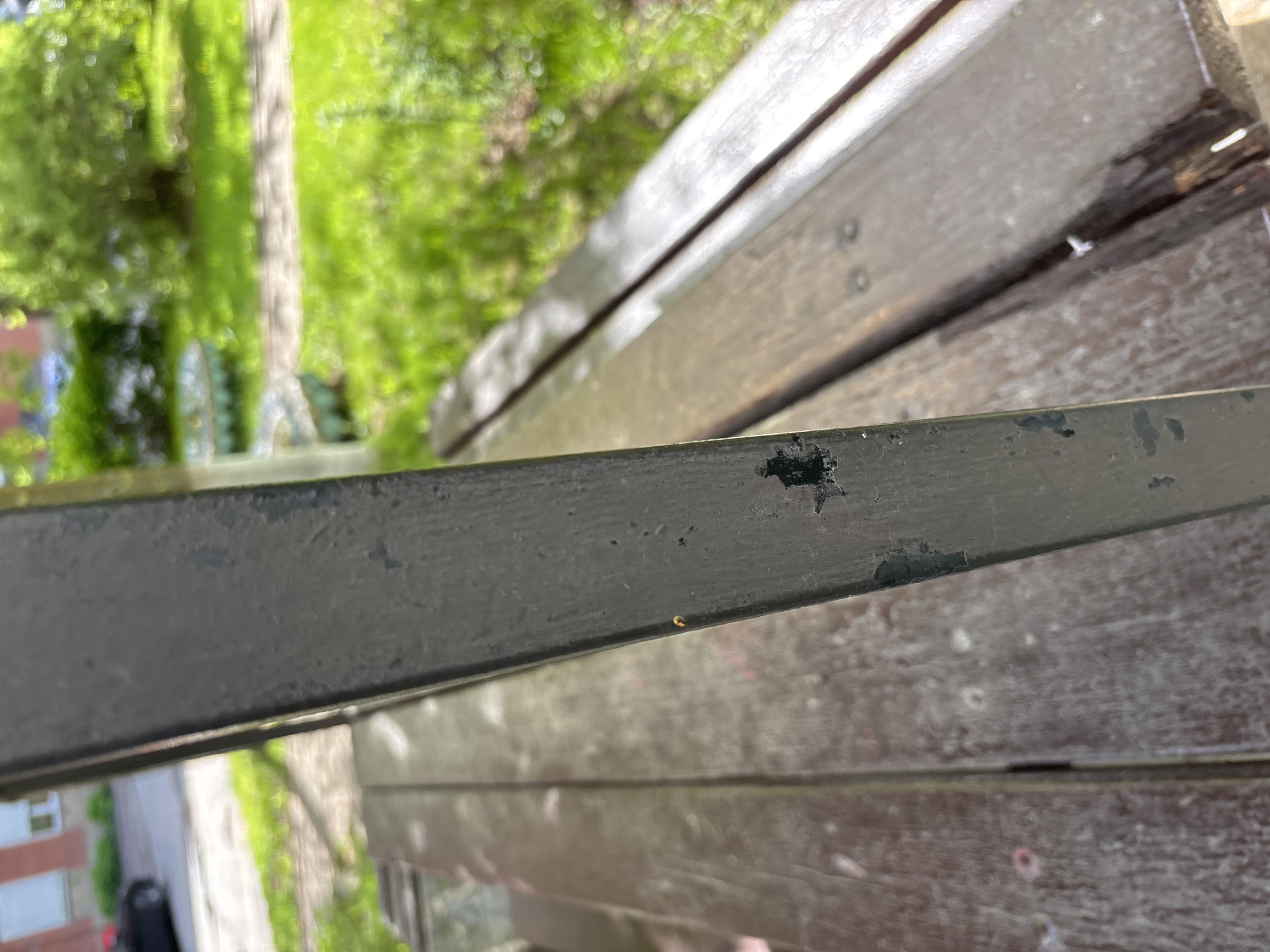 The image shows a close-up, angled view of a weathered park bench, likely in a public space in Saint Petersburg, Russia. The focus is on a section of the metal armrest, which is painted a dark gray or black. The paint is chipped and peeling in several areas, revealing the underlying material, suggesting age and exposure to the elements. Behind the armrest are wooden slats that form the seating area of the bench. The wood is also weathered and appears to be stained or painted a dark brown. In the background, out of focus, are green trees and some buildings, indicating an urban park or garden setting. The time of day appears to be daytime, based on the natural light in the scene. There are no people visible in the image.