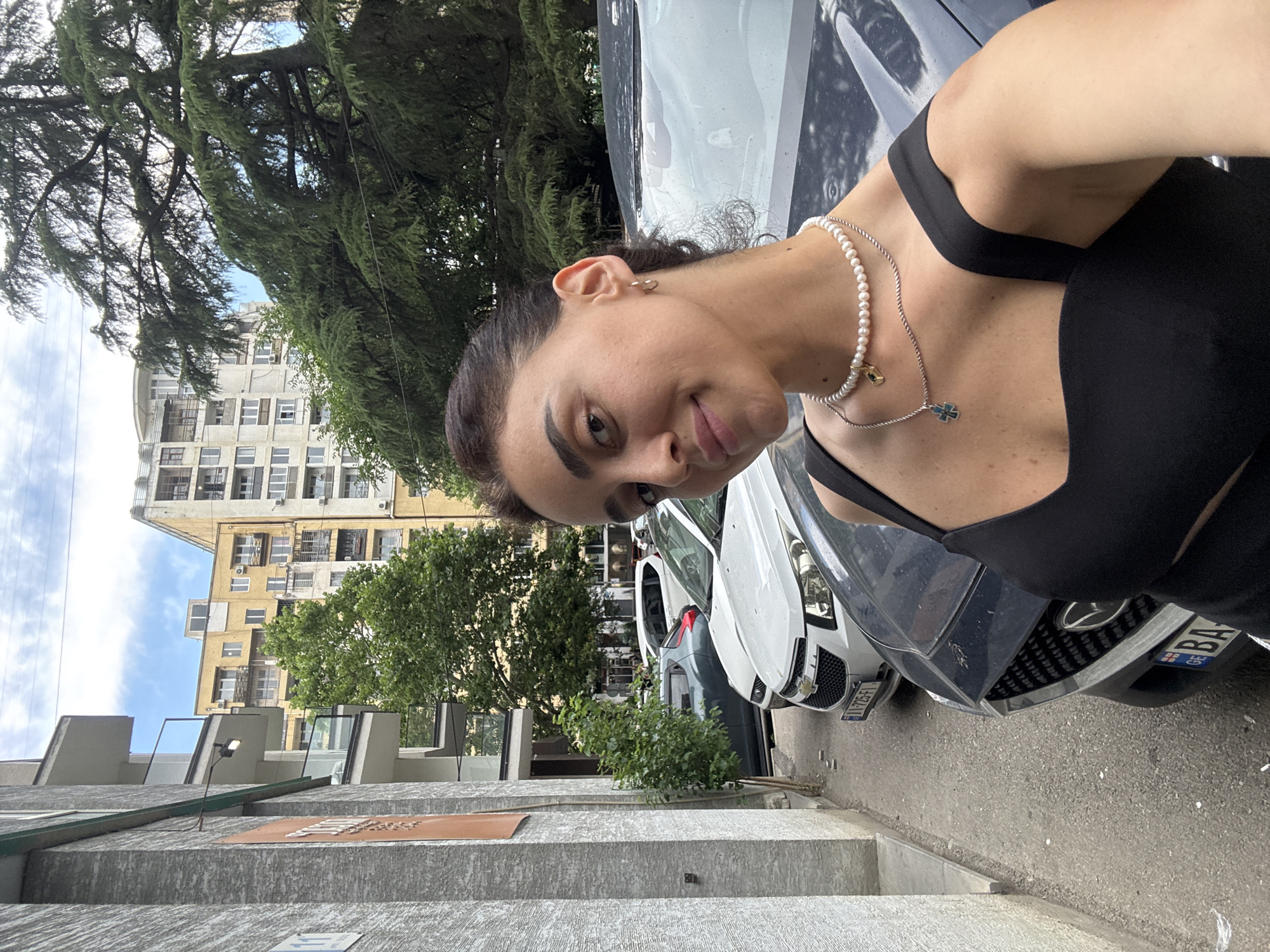 A woman takes a selfie in front of parked cars on a street in Tbilisi, Georgia. The woman is wearing a black dress and a pearl necklace. The background shows apartment buildings, trees, and cars.