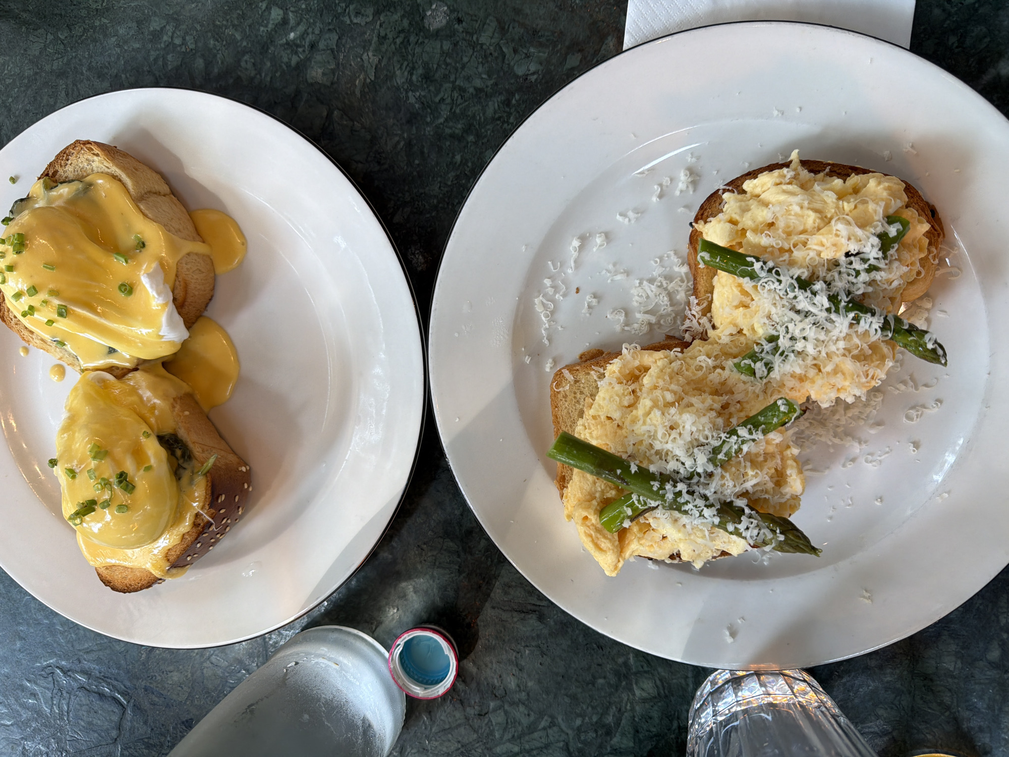 Two plates of food sit on a dark green, possibly marble, surface. The plate on the left holds two pieces of bread topped with what appears to be eggs benedict, covered in a yellow sauce and garnished with green chives. The bread seems to be a sesame seed roll sliced in half. The plate on the right contains two pieces of toast topped with scrambled eggs, asparagus spears, and grated cheese. The plates are white with black trim. Below the plates are parts of a water bottle and a glass. This is a still life photo of a breakfast or brunch meal, possibly taken at a restaurant or cafe. The scene lacks other details suggestive of a specific location in Tel-Aviv, Israel.