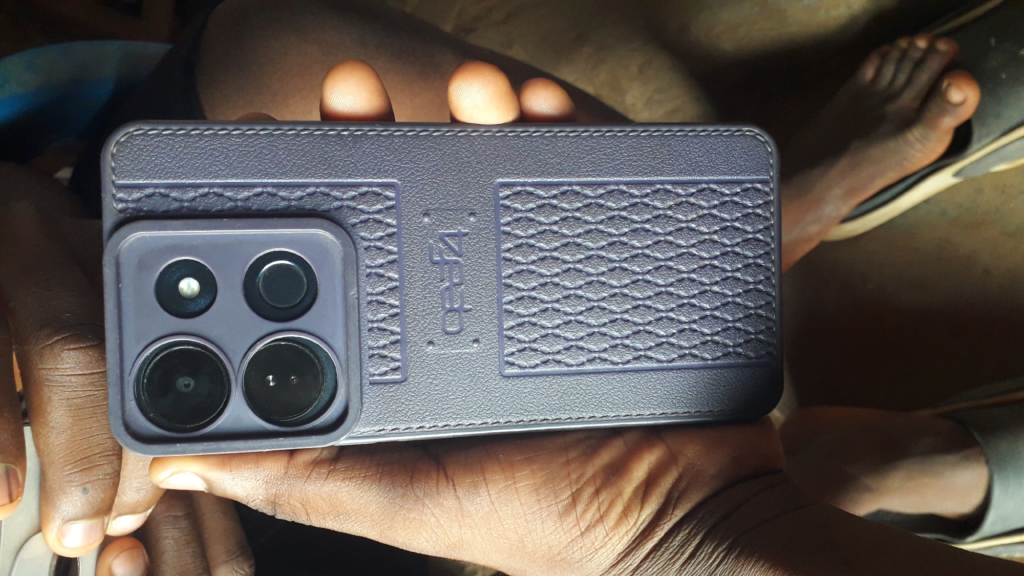 A person is holding a purple cell phone in their hands. The phone has a case with a textured design and four camera lenses arranged in a square. The photo was taken in Nkpor, Nigeria. The subject is a person of African descent. A foot can be seen in the background, partially obscured by a slipper.