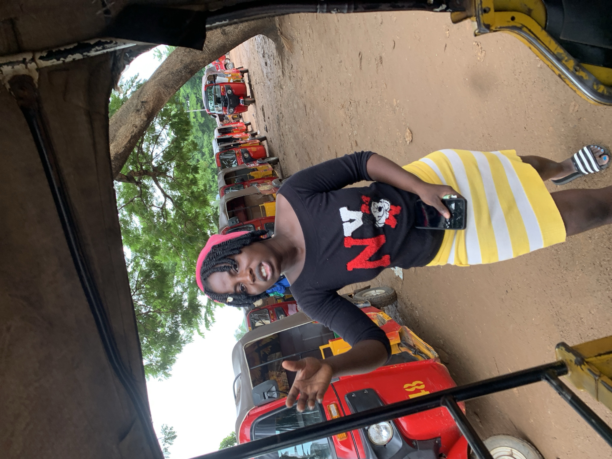 The image shows a young woman with braided hair and a pink headband standing outdoors, in what appears to be a marketplace or transport hub. She is wearing a black long-sleeved shirt with a red and white graphic design on it, and a yellow and white striped skirt. She is holding a smartphone in her right hand. Her facial expression suggests she is speaking or reacting to something, with an open mouth and raised eyebrows.
Behind her, a line of red and yellow auto-rickshaws, also known as "Keke Napep" or "Marwa," are parked on a dirt road. These vehicles are a common mode of public transportation in Nigeria. The background shows lush green trees and foliage, indicating an outdoor, possibly tropical, setting. The lighting suggests it is daytime, with overcast skies. The overall scene is vibrant and full of local character, with the parked vehicles and the woman's attire hinting at a busy commercial or transportation area in Abuja, Nigeria. There is some visible text on one of the auto-rickshaws, "81", in yellow lettering.