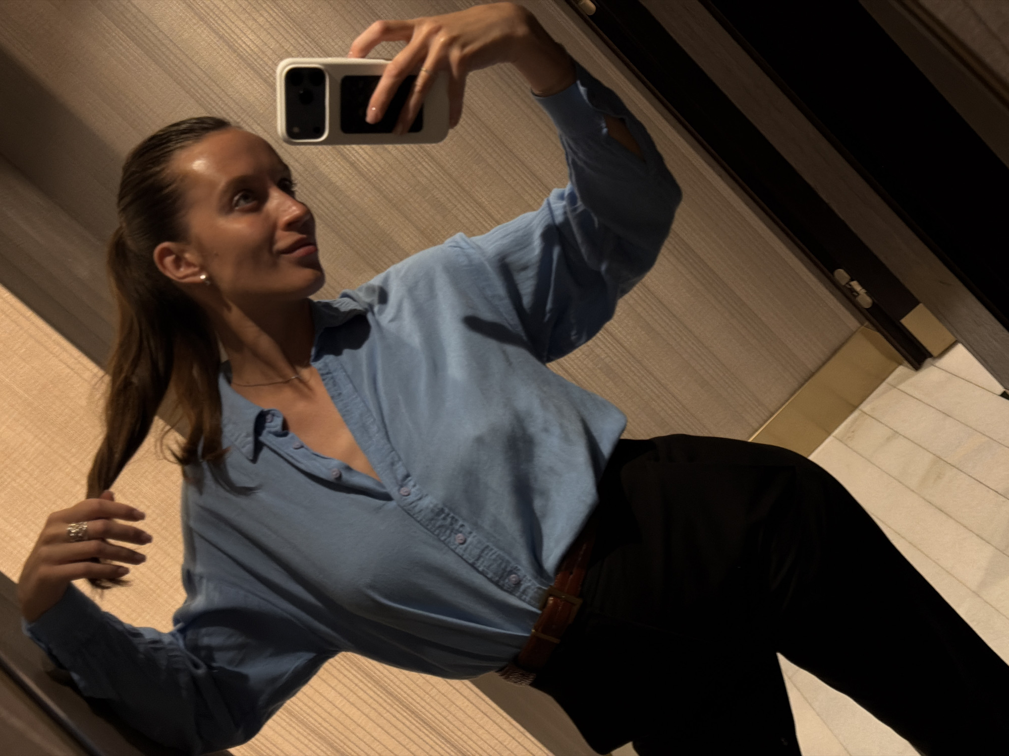 A young woman is captured in a mirror selfie, standing indoors in what appears to be a room or hallway in Budva, Montenegro. She holds a white smartphone with a dark camera module in her raised right hand, seemingly taking the photograph. Her left hand gently holds a section of her dark hair, which is styled in a ponytail. She wears a light blue, long-sleeved button-up shirt and black trousers, cinched with a brown belt featuring a metallic buckle. A delicate necklace, an earring, and a ring on her left hand are also visible.
The woman looks upwards and to her right with a soft, slightly smiling, and relaxed expression. Her skin appears tan. The background features light brown or beige walls with a subtle horizontal textured pattern, possibly wood paneling or wallpaper. Dark vertical elements, perhaps door frames or furniture edges, are visible on the right. The flooring, visible in some areas, appears to be light-colored tiles. The lighting suggests an indoor setting, possibly with artificial light, creating a slightly muted but comfortable atmosphere. No text is visible in the image.