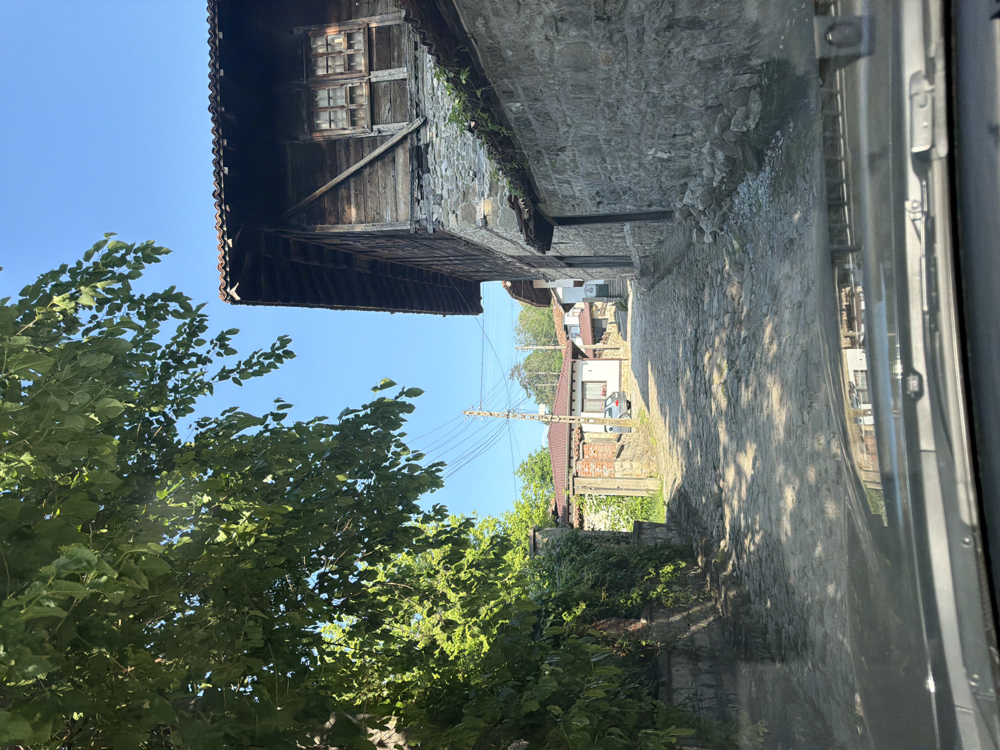 This image, captured from within a vehicle as indicated by the visible dashboard and windshield reflections at the bottom, displays a cobblestone street ascending through Elena, Bulgaria, under a clear blue sky. Dense green foliage from an overhead tree occupies the upper left quadrant, casting shadows across the uneven cobblestone path. On the left, a stone wall is heavily covered in lush green vines. Further up the incline, a house with a red-tiled roof and white facade is partially visible on the left. On the right side of the street, a large, traditional structure features a stone foundation topped by an upper story constructed from dark, weathered timber with prominent window frames containing multiple small panes, all beneath a red-tiled roof. A contiguous stone wall borders the right side of the street. Beyond this, another building with white walls and a red-tiled roof is present, with a silver car parked in front. A utility pole with multiple power lines stands along the street further into the distance. No people, specific actions, or interactions are visible in the scene.
