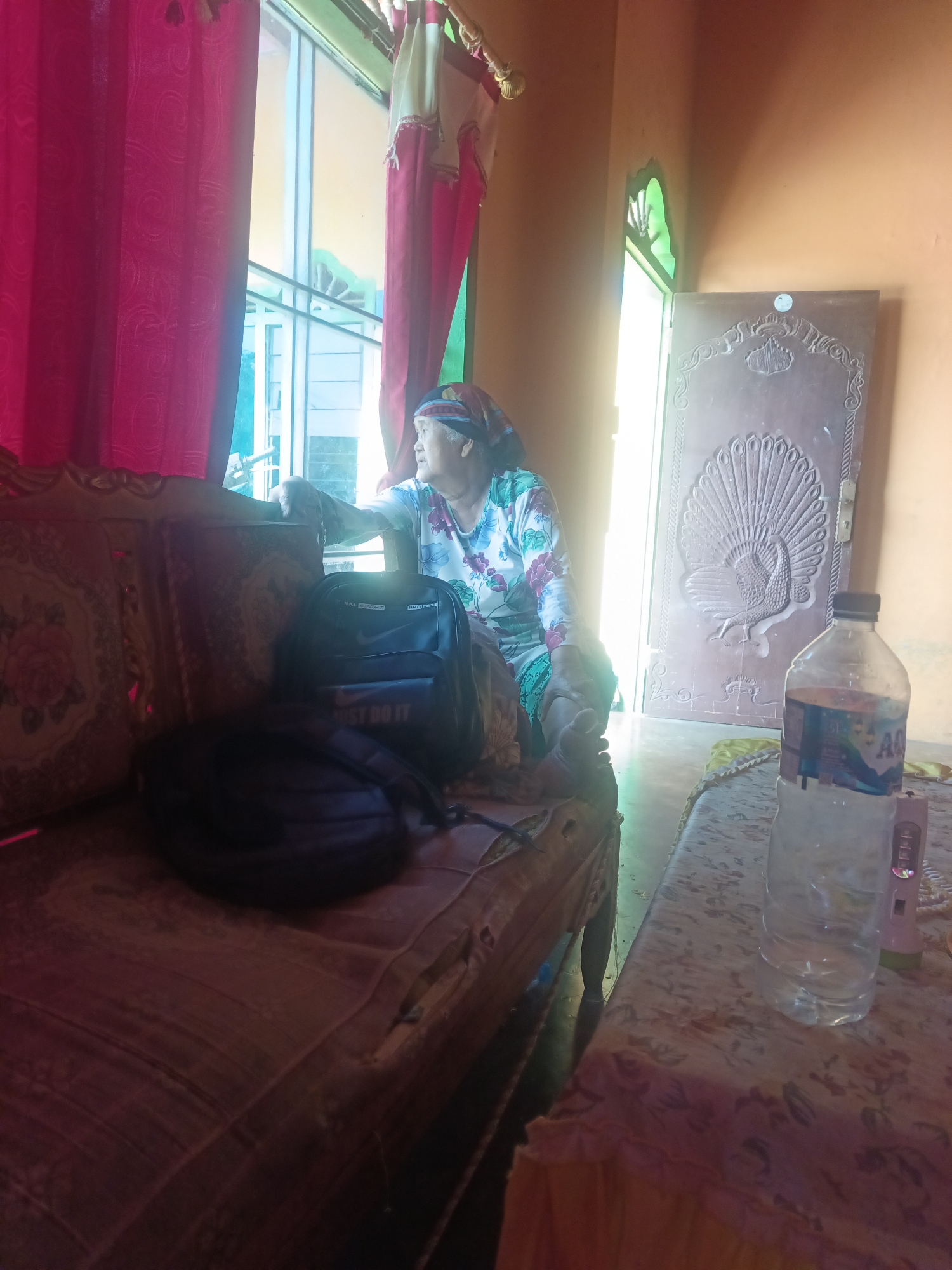 An outdoor scene in Terbanggi Marga, Indonesia, features a woman sitting on a couch by a window. She is wearing a headscarf and a floral print shirt. In front of her, on the couch, are two backpacks, one black and one dark. A partially filled plastic bottle of water and a pink electronic device are on a table to the right. The room has orange walls and a wooden door with carved designs. The lighting suggests it is daytime, with sunlight streaming in from the window and door.