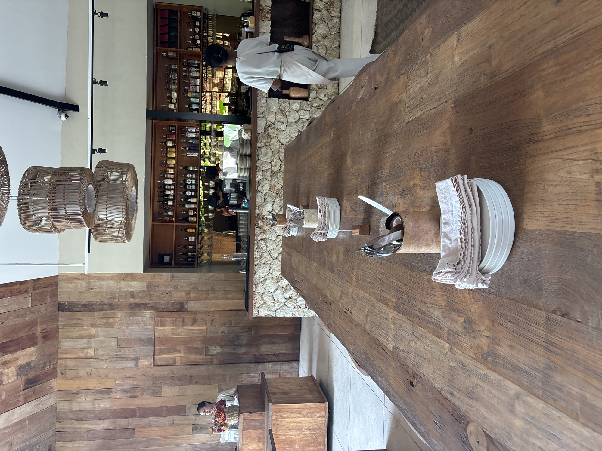 This image depicts the interior of a restaurant or bar, showcasing a rustic and natural aesthetic. In the foreground, a substantial, dark wooden table with visible grain and a live edge runs across the frame. On this table, stacks of white plates are neatly arranged beside crumpled linen napkins, and several decorative holders, possibly for cutlery, are present.
In the mid-ground, a bar area is visible, featuring shelves well-stocked with various bottles of liquor and wine. The base of the bar counter is clad with rough, light-colored stone. A staff member, dressed in a white uniform, stands behind this bar, appearing to be at work.
To the left, three large, circular light fixtures crafted from woven rattan hang from the ceiling. The left wall is covered in horizontally laid wood paneling of various shades. In the lower-left corner, another individual is partially visible, holding a basket filled with flowers and looking towards the camera.
The ceiling is white, equipped with a black track holding several spotlights and a small security camera. The floor is covered in light-colored tiles. The overall setting suggests a welcoming, possibly upscale, establishment. The scene appears to be taking place during the day, illuminated by indoor lighting. No visible text is clearly discernible. The décor aligns with the provided context of Kuta Selatan, Indonesia.