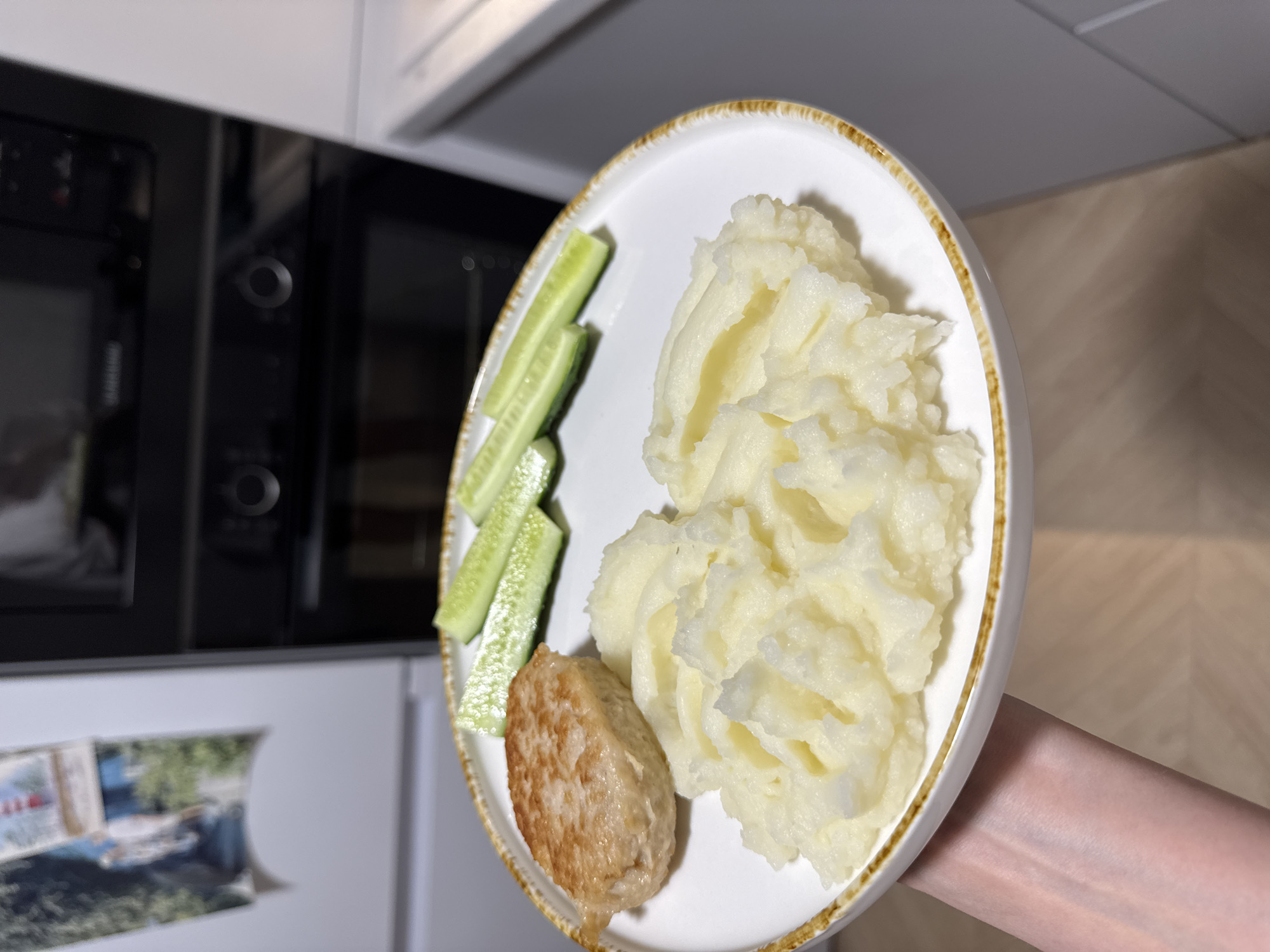 In the city of Postol, Russia, a white plate with a brown rim holds a meal. The plate is positioned on a light-colored countertop. On the plate, there is a golden-brown patty on the left, and a serving of fluffy mashed potatoes to the right and front. Three sliced cucumbers are arranged in a row behind the patty. The background is blurred, showing a modern kitchen environment with kitchen appliances. The image is taken from a slightly elevated angle, with a hand holding the plate visible in the lower left corner.