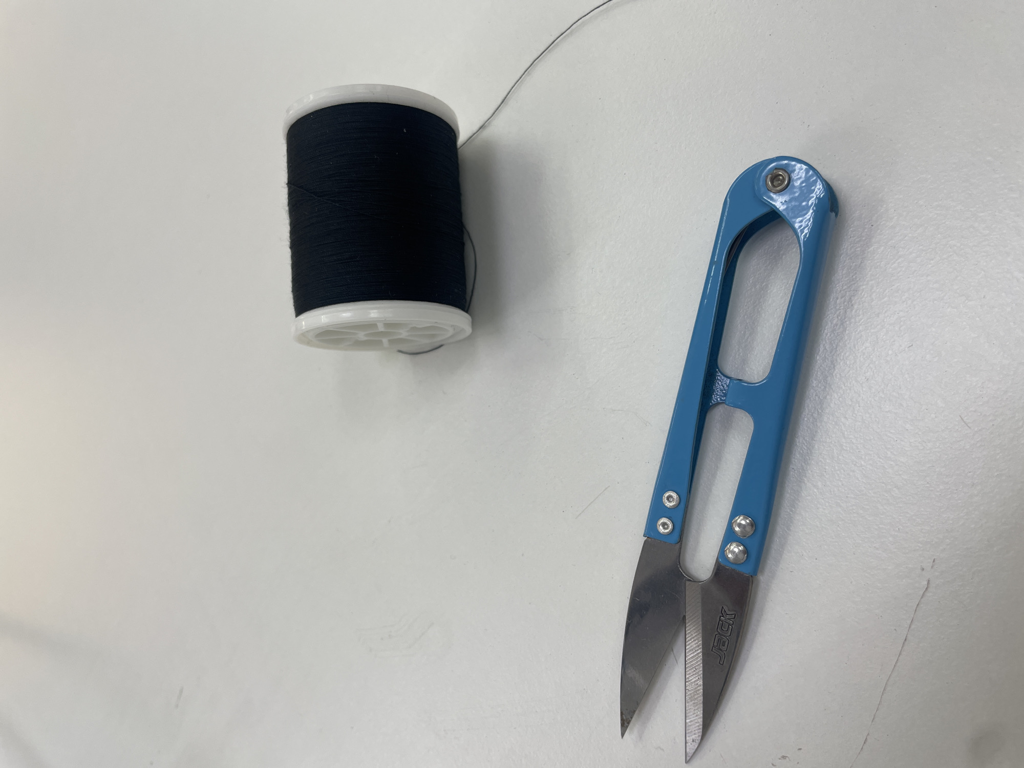 The image displays a spool of black thread and a pair of blue thread snips resting on a white, textured surface. The thread snips are positioned in the lower left quadrant of the image, with the blades open and the handle forming a loop. The spool of thread is situated in the upper right quadrant, with a single strand of thread extending from it. The lighting is bright and overhead, casting subtle shadows on the surface. The background is a plain white expanse, suggesting an indoor setting. There are no people or animals visible in the image. The location is not specified within the image.