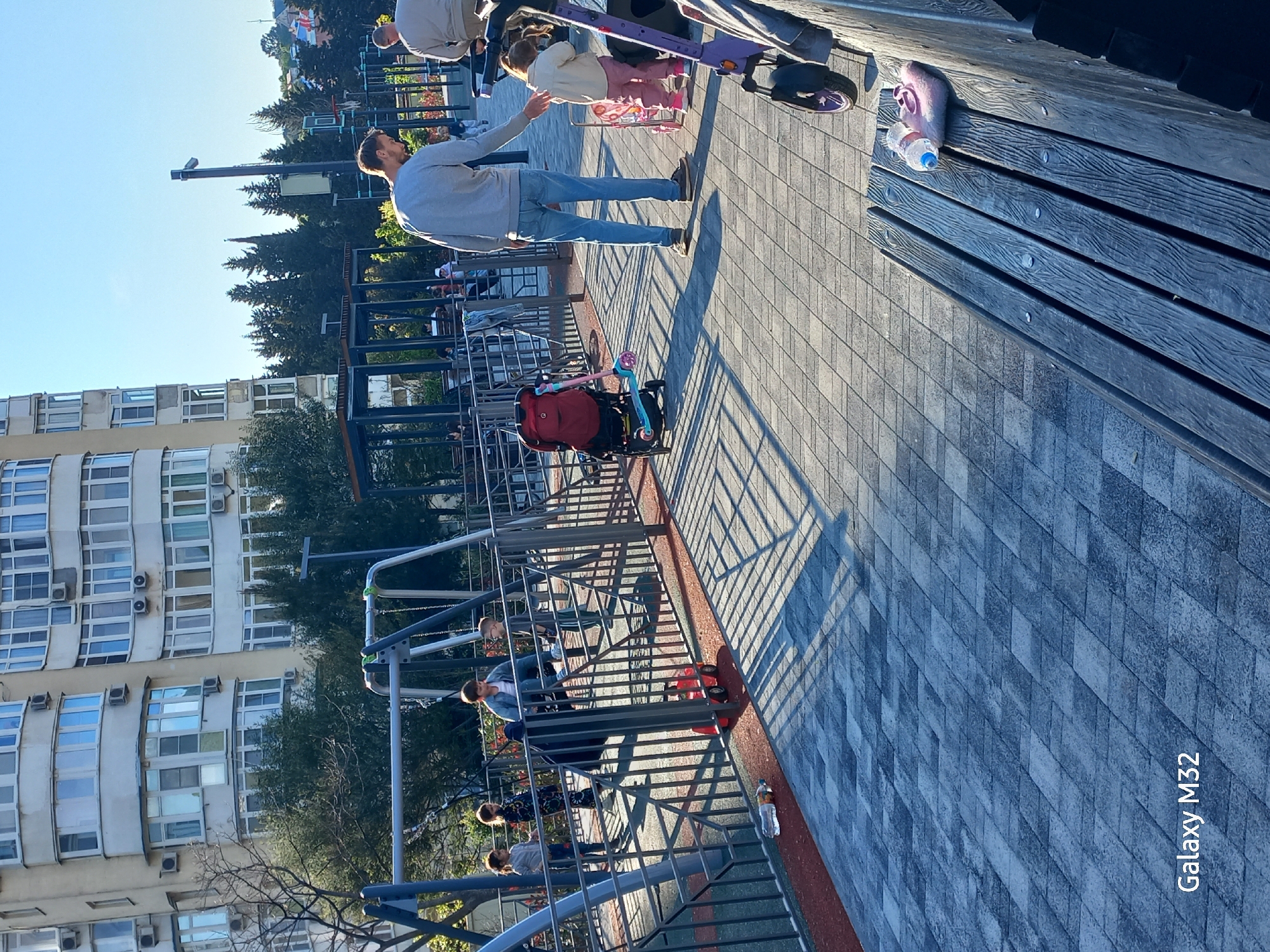 The image shows an outdoor scene on a sunny day, likely in an urban area. A man wearing a gray shirt and jeans stands on a paved walkway, appearing to interact with a child on a scooter. There is another adult nearby. To the left of the walkway, there's a playground with swings and a fence. A red stroller is parked next to the fence. Further in the background, a large building with many windows is visible, indicating an apartment complex. Behind the building, there are trees and foliage. A wooden plank is present in the frame. The picture orientation makes the objects and people appear tilted. The inscription "Galaxy M32" is in the lower right corner.