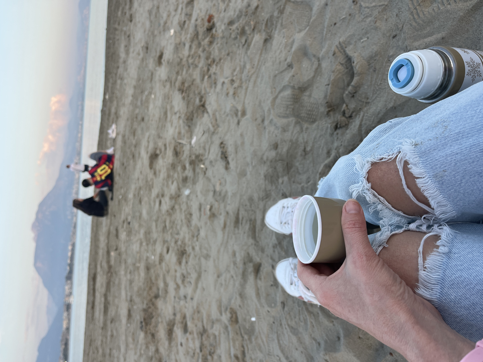 The image depicts a person sitting on a sandy beach with mountains and water visible in the background. The person is wearing ripped light blue jeans and white sneakers. They are holding a tan-colored cup in their right hand. A white and silver thermos with a blue top is positioned near their leg. In the distance, there is a small group of people on the beach. The location is likely Alanya, Turkey, as mountains and the sea are visible. The scene suggests a casual and relaxed atmosphere on the beach.