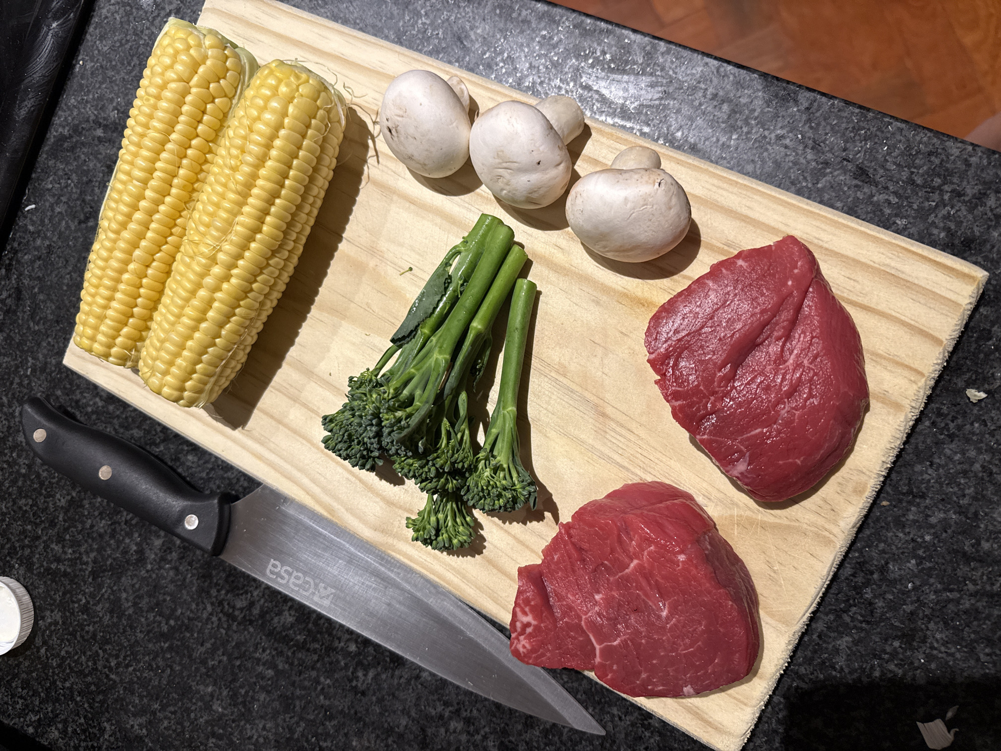 A wooden cutting board sits on a dark countertop. Arranged on the board are two ears of corn, three white mushrooms, a bunch of broccolini, and two cuts of raw red meat. A kitchen knife with a black handle lies on the countertop next to the cutting board. The setting does not explicitly reveal its location within Cape Town, South Africa. No people are visible in the image.