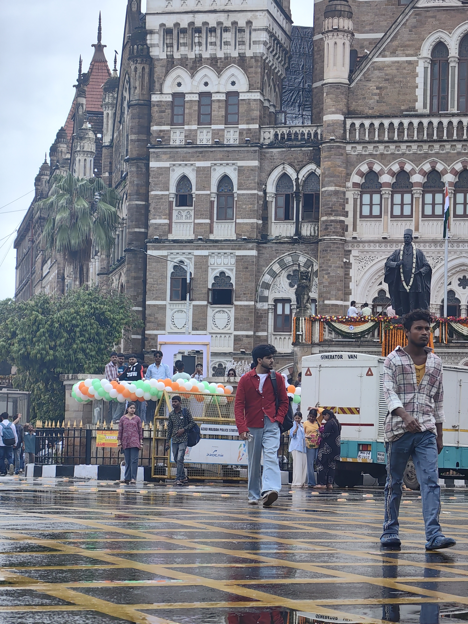 The image depicts a street scene in Mumbai, India, in front of the Chhatrapati Shivaji Maharaj Terminus, formerly known as Victoria Terminus. The iconic railway station, a UNESCO World Heritage Site, serves as a grand backdrop with its intricate Victorian Gothic architecture. In the foreground, several people are walking across a wet street, indicated by reflections on the asphalt. The street is marked with yellow lines, suggesting it's a pedestrian crossing or traffic lane.
On the right side of the image, a young man in a plaid shirt and ripped jeans walks towards the viewer. To his left, another man in a red shirt and light blue jeans walks in the opposite direction. In the middle ground, a group of people are gathered near a white generator van. Some individuals are standing behind a barrier adorned with balloons in the colors of the Indian flag (orange, white, and green). A statue of a distinguished figure, likely a historical personality, stands prominently on a platform in front of the station, decorated with floral garlands. The sky is overcast, and the wet pavement suggests recent rain. The overall atmosphere is that of a public space, possibly during an event or a busy day.