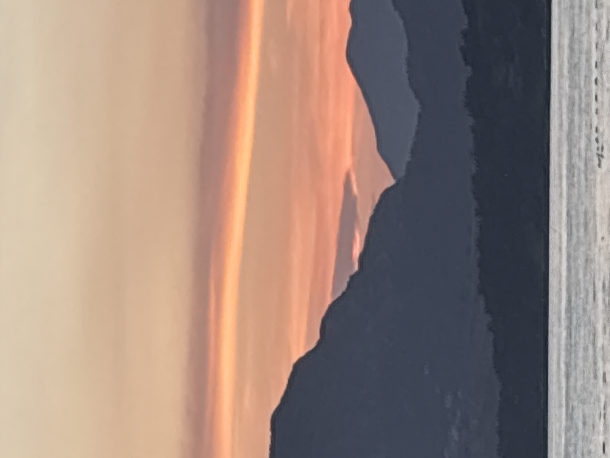 The image depicts a serene coastal landscape at dusk. In the foreground, the calm, reflective surface of the Adriatic Sea stretches across the bottom of the frame. A thin band of what appears to be a buoys or markers stretches horizontally across the lower right portion of the sea.
Dominating the mid-ground are the dark, silhouetted forms of rolling hills and mountains. These landmasses are densely covered with trees, creating a textured, dark mass against the sky. The contours of the mountains suggest a rugged, natural topography.
The sky above is a spectacle of warm hues characteristic of a sunset. A gradient of colors ranges from a soft, peachy-orange near the horizon to a pale, creamy yellow at the top of the frame. A distinct horizontal band of vibrant orange cuts across the sky, likely indicating a cloud formation catching the last rays of the sun.
The scene is devoid of any human presence or explicit man-made structures, emphasizing the natural beauty of the environment. The overall impression is one of tranquility and the fading light of day over the coastal region of Bar, Montenegro.
