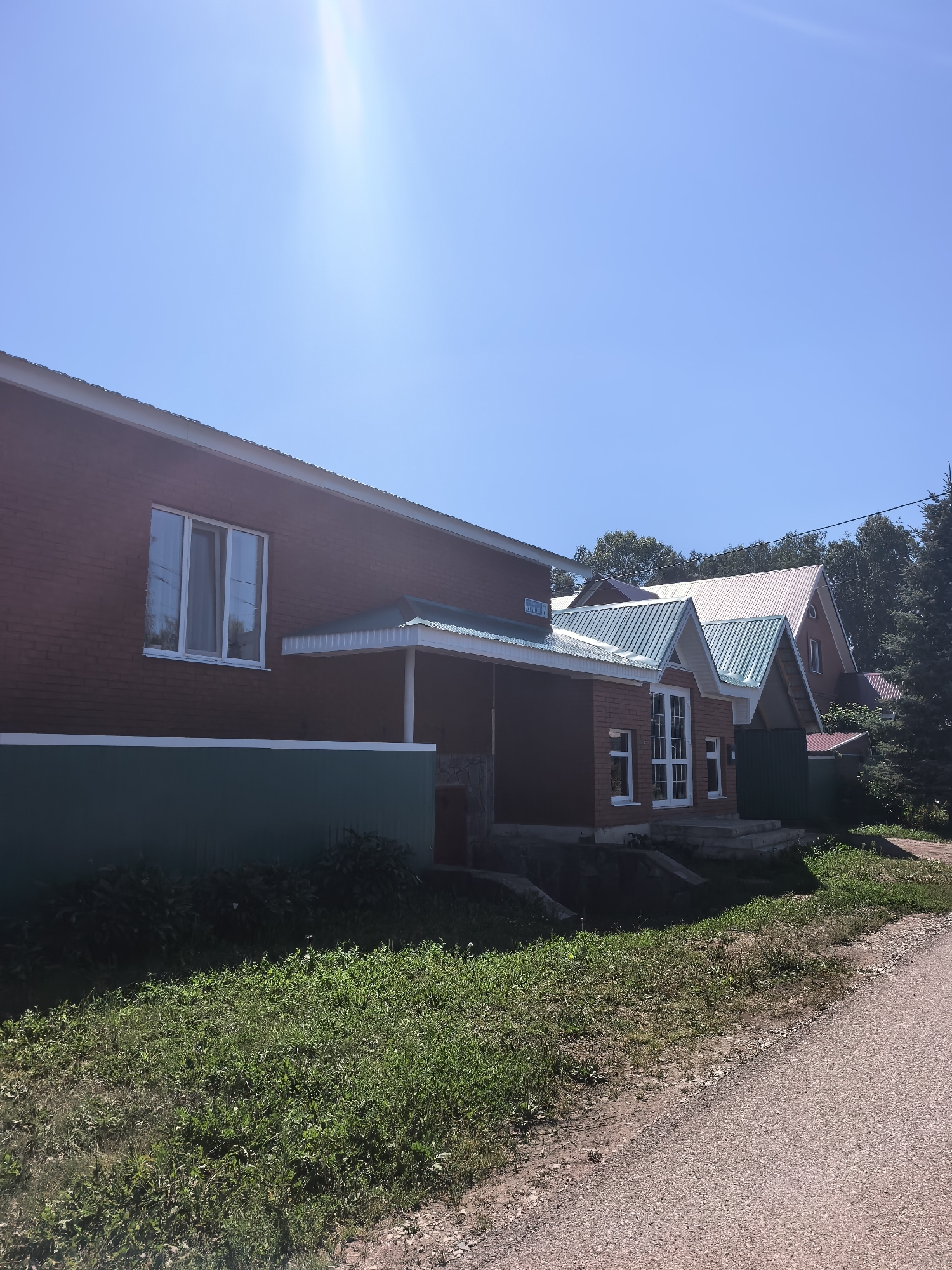 This image depicts a sunny day scene in Sterlitamak, Russia, featuring a row of modern brick houses. The sky is a clear, bright blue, with a lens flare from the sun casting a bright ray across the upper left portion of the frame, indicating mid-day. The houses have distinctive green metal roofs with white trim, and some feature prominent gabled sections.
The house on the left is a single-story structure with a large, white-framed window. A green wainscoting runs along the base of this house, contrasting with the red brick. To the right, a slightly recessed porch area with a green roof is visible, supported by white columns. Further to the right, a larger, multi-story house with a light-colored gabled roof is partially visible, with trees lining the background.
In front of the houses, there is a strip of green grass and some smaller plants, leading towards a gravel road on the right side of the image. The overall setting suggests a residential area with newly constructed or well-maintained homes. The atmosphere appears peaceful and quiet, typical of a suburban or rural environment on a clear day. No people are visible in the image, and no discernible text is present. The time of day is clearly daytime, and the weather is sunny and fair.