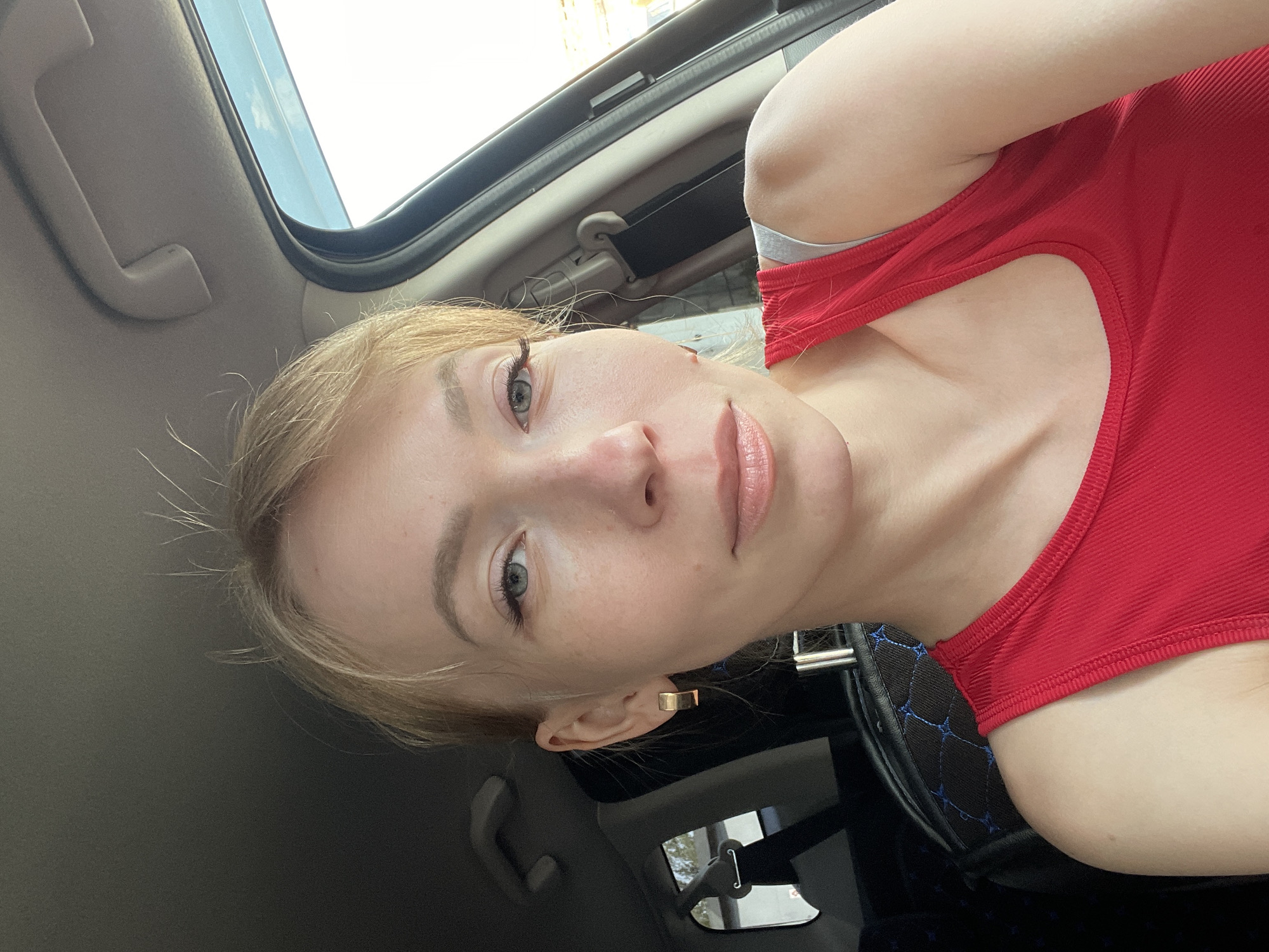 A light-skinned woman with blonde hair and light eyes is sitting in a car. She is wearing a red tank top. The interior of the car includes a sunroof, a steering wheel with a blue stitched cover, and a seatbelt. The image was taken from a low angle, looking up at the woman. The location is likely Perm, Russia.