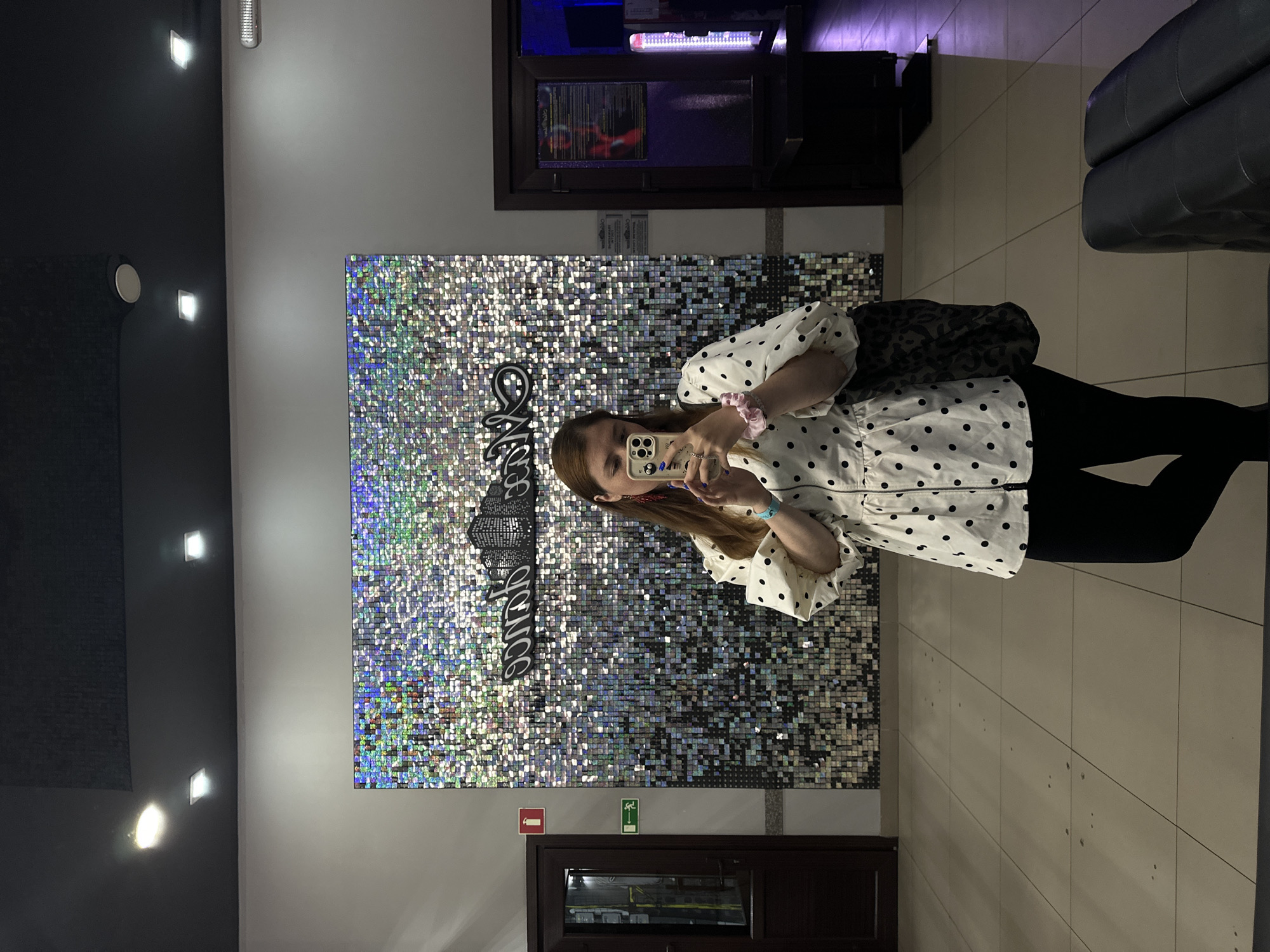 A woman with reddish-brown hair is standing indoors and taking a mirror selfie with her phone. She is wearing a white blouse with black polka dots and puffed sleeves, and black leggings. She has a pink scrunchie on her wrist and red, dangly earrings. Her phone case is light yellow and has stickers on it. She is carrying a black bag with a leopard print strap. Behind her is a wall covered in small, reflective square tiles, creating a shimmering effect. A stylized black sign with building silhouettes is also visible on the wall. To her left is a dark brown door with a red fire extinguisher sign and a green exit sign above it. To her right is another doorway leading to a dimly lit area with purple and blue lights. The floor is tiled in a beige, square pattern. The overall environment appears to be an entertainment venue or establishment in Petropavlovsk-Kamchatsky, Russia.