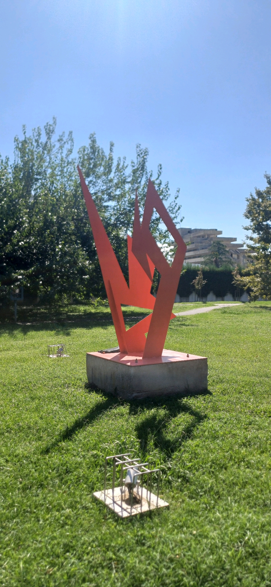 The image depicts an outdoor scene under a clear blue sky. In the foreground, a bright orange abstract sculpture is mounted on a grey concrete base. The sculpture is composed of sharp, angular metal planes. In front of the main sculpture, on the green grass, is a smaller, cage-like metal structure containing a grey object. The area surrounding the sculptures is a manicured lawn with lush green grass. In the background, several trees with dense green foliage are visible, and beyond them, a tiered, modern building with horizontal lines is situated. A paved pathway winds through the grass in the middle ground. The image appears to have been taken during daylight, with sunlight casting shadows on the grass. There are no people visible in the scene. The setting is likely a park or public garden within the city of Abano Terme, Italy, given the architectural style of the background building, which resembles elements of the 'Palazzo Montirone' or a similar modern structure in the area.