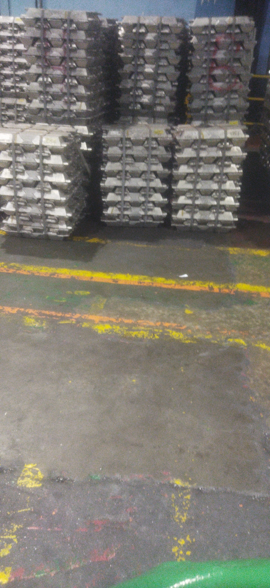 The image depicts stacks of aluminum ingots in what appears to be a warehouse or industrial facility. The ingots are silver-colored and are arranged in neat vertical columns and horizontal layers, secured by metal strapping. In the foreground, the concrete floor is marked with yellow and orange lines, possibly indicating safety zones or pathways. The lighting suggests an indoor environment, with some areas illuminated and others in shadow. There are no people or specific landmarks to identify the location within Cileungsi, Indonesia.