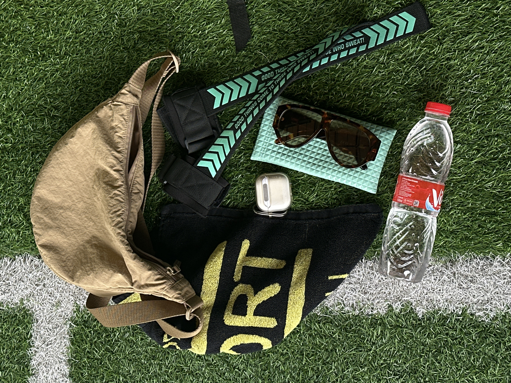 This is a high-angle, flat lay photograph of workout gear arranged on artificial turf, likely a sports field, marked with a white line at the top left. The items are placed on a green, artificial grass surface.
From top to bottom and left to right, the objects include: a partially empty clear plastic water bottle with a red cap, lying on its side; a black towel with yellow lettering that appears to spell out "SPORT"; a small, rectangular, white, metallic container; a light green textured rectangular case; a pair of tortoiseshell sunglasses resting on the case; a tan-colored hip pack or fanny pack with a zippered main compartment and a strap; and a pair of black ankle or wrist straps with teal chevron patterns and white text that reads "GOOD THINGS COME TO THOSE WHO SWEAT!".
The overall scene suggests preparation for or aftermath of a workout. The environment is a sports field, indicated by the artificial turf and white line. The specific location in North Kuta, Indonesia, is not evident from the visual information in the image. There are no people or actions depicted.