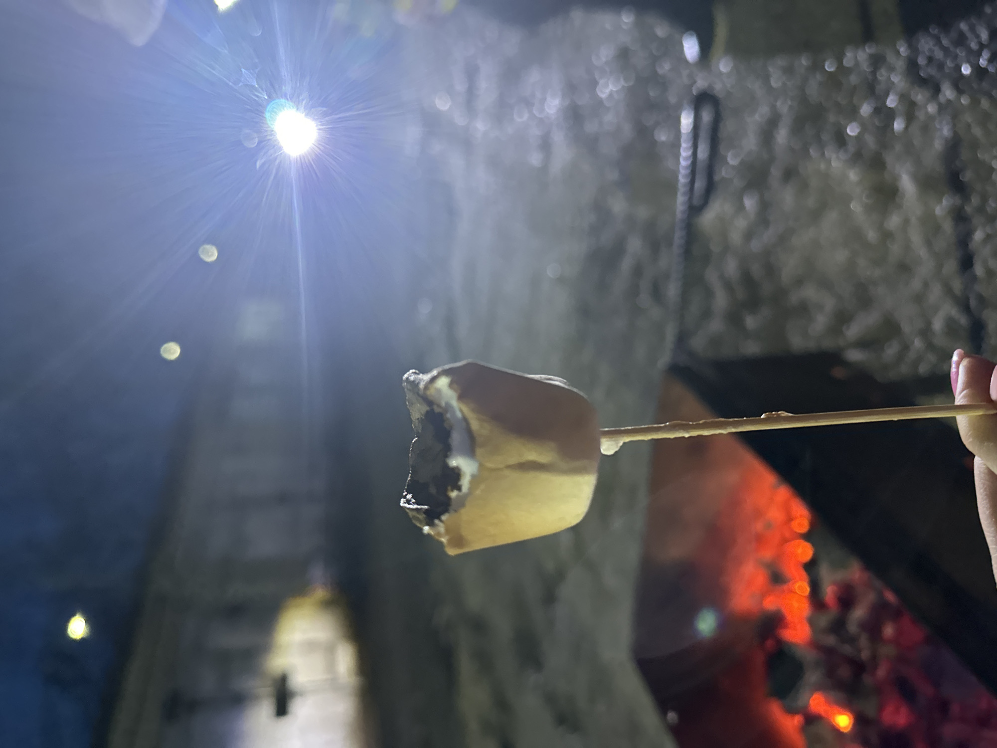The image captures a close-up of a partially toasted marshmallow on a wooden skewer, held by a person's hand in the lower right. The marshmallow is light brown, with some darker, possibly burnt or eaten, sections. Directly below and to the right of the skewer, a fire pit with glowing orange embers provides warmth and light, casting a reddish-orange hue.
The background is dark and features textured surfaces, resembling rough stone, rock, or concrete walls, possibly in a cave, grotto, or an enclosed underground space. These surfaces appear somewhat damp or reflective, creating soft, out-of-focus bokeh effects from various light sources. In the upper left portion of the image, a very bright, starburst-like artificial light source is prominent, casting a strong lens flare across the dark upper-left quadrant. Several other smaller, diffused circular light sources are scattered throughout the darker background, contributing to the cavernous and dimly lit atmosphere.
The activity depicted is the roasting or consumption of a marshmallow, suggesting a recreational or social event around a fire. The scene appears to take place at night or in an environment without natural light. There are no visible textual elements in the image.