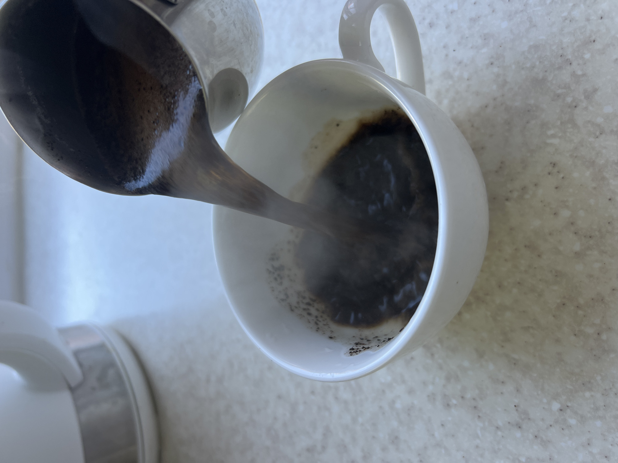 A close-up, overhead shot shows dark, hot coffee being poured from a metal pot into a white ceramic mug. Steam rises from the liquid as it fills the mug, which sits on a speckled, light-colored countertop. In the background, the base of a white electric kettle is visible. The scene is brightly lit, suggesting it is daytime, and there is no indication of a specific location within Fontanka, Ukraine.