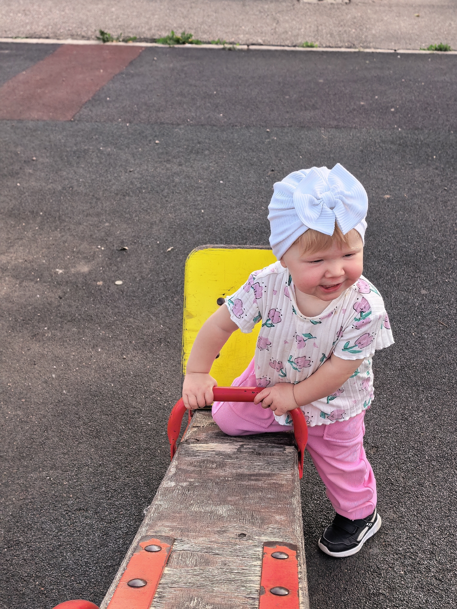 A Caucasian toddler with blond hair and a white bow headband is on a playground seesaw. She is wearing a white t-shirt with pink floral patterns and pink pants. Her left leg is bent and resting on the seesaw, while her right foot is on the ground. She is holding onto the red handle of the seesaw. The playground surface is dark asphalt with some red and gray patches. A yellow metal structure is visible behind her. The image was taken outdoors in Sterlitamak, Russia.
