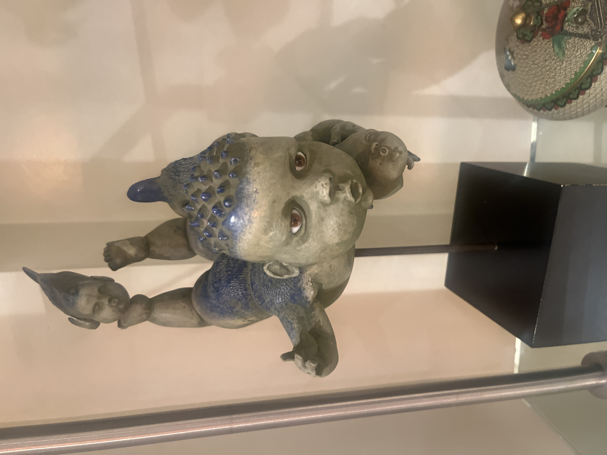 A ceramic sculpture of a fantastical, baby-like figure is centrally displayed on a clear glass shelf. The main figure is rendered in pale green-grey, featuring large brown eyes and pursed lips. Its torso and crown are adorned with dark blue, textured, spiky protrusions. The figure's left arm is raised, with the thumb extended upwards. Two additional smaller, full baby-like heads are attached to the main figure: one to its lower left limb and another positioned beneath its main head. The sculpture is mounted on a slender dark metallic rod, which is inserted into a black rectangular base. The background consists of a plain, light-colored wall. A vertical metallic pole is visible along the left edge of the frame. A partially visible spherical object, exhibiting an intricate pattern, a gold finial, and red floral motifs, is located on the shelf to the right. The scene is located in Mexico City, Mexico.