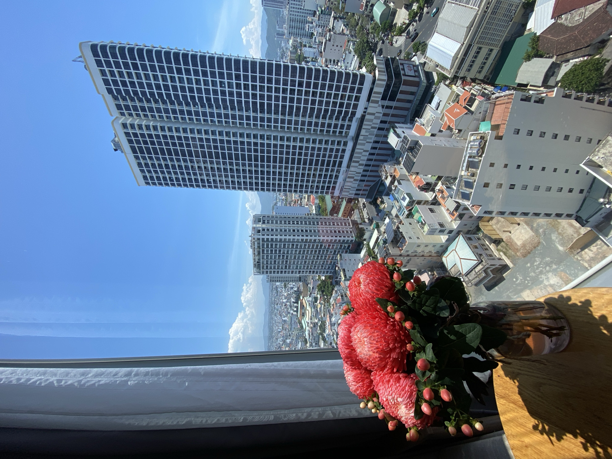 The image shows a high-angle view of a city with a bouquet of pink chrysanthemums and small red berries in a glass vase placed on a wooden table in the foreground. The flowers and vase are on the left side of the frame, casting a shadow on the table. Beyond the flowers and table, a large window looks out onto a cityscape.
The city appears to be densely populated with numerous buildings of varying heights and architectural styles. Two prominent, tall, modern white buildings with dark grid-like window patterns dominate the central and right portions of the background. There are also numerous smaller residential and commercial buildings, some with red-tiled roofs. The city extends towards a distant mountainous landscape under a clear blue sky with scattered white clouds. The overall impression is an urban environment, likely in a coastal city, given the distant mountains and the general atmosphere. The location is identified as Nha Trang Ward, Vietnam.
