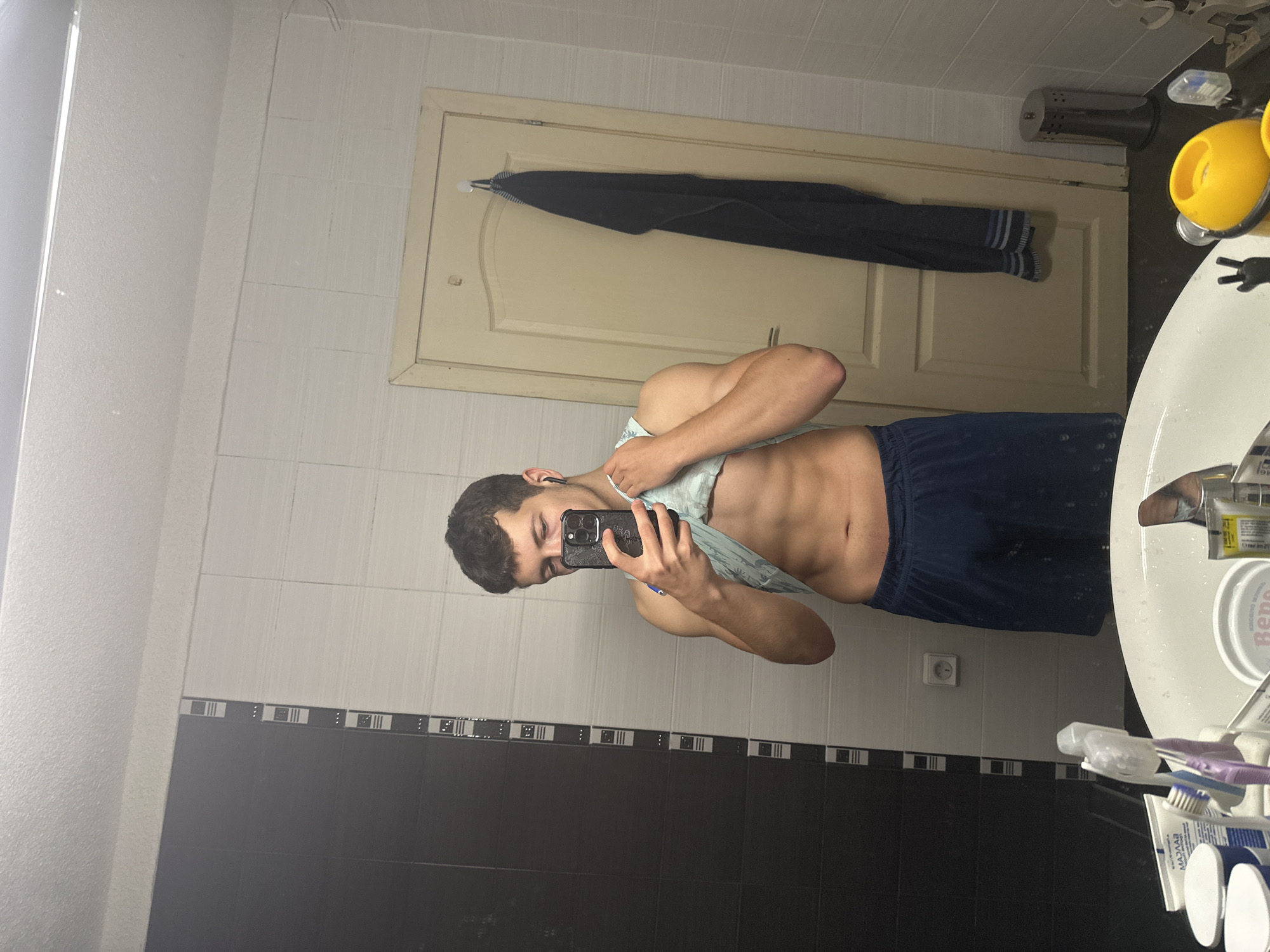 A young man with dark hair and fair skin is standing in a bathroom and taking a mirror selfie. He is wearing blue athletic shorts and a light blue tank top that he has lifted to reveal his abdomen. He is holding a black smartphone in his right hand, with which he is taking the picture. The background shows a bathroom with white tiled walls, a black and white tiled accent wall, a light beige door, and a white sink. A dark blue towel is hanging on the door. There are various toiletries on the sink and on a shelf. The lighting is dim, suggesting it is nighttime or the lights are low.