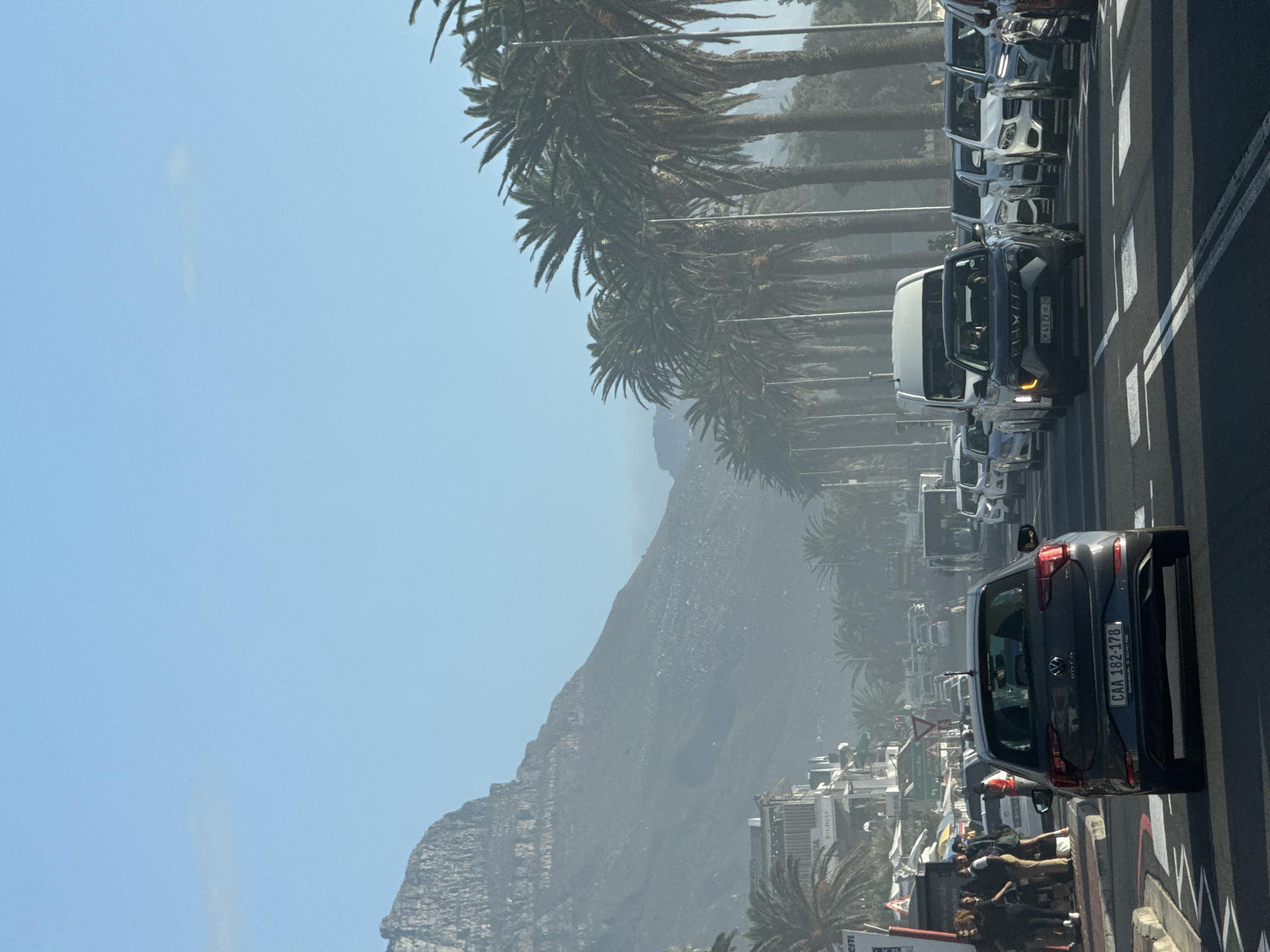 The image shows a street scene in Cape Town, South Africa. In the background is a hazy view of Lion's Head mountain. Palm trees line the street, which is busy with traffic. A dark grey Volkswagen Polo with the license plate CAA 182-178 is in the foreground. A line of pedestrians is on the sidewalk.