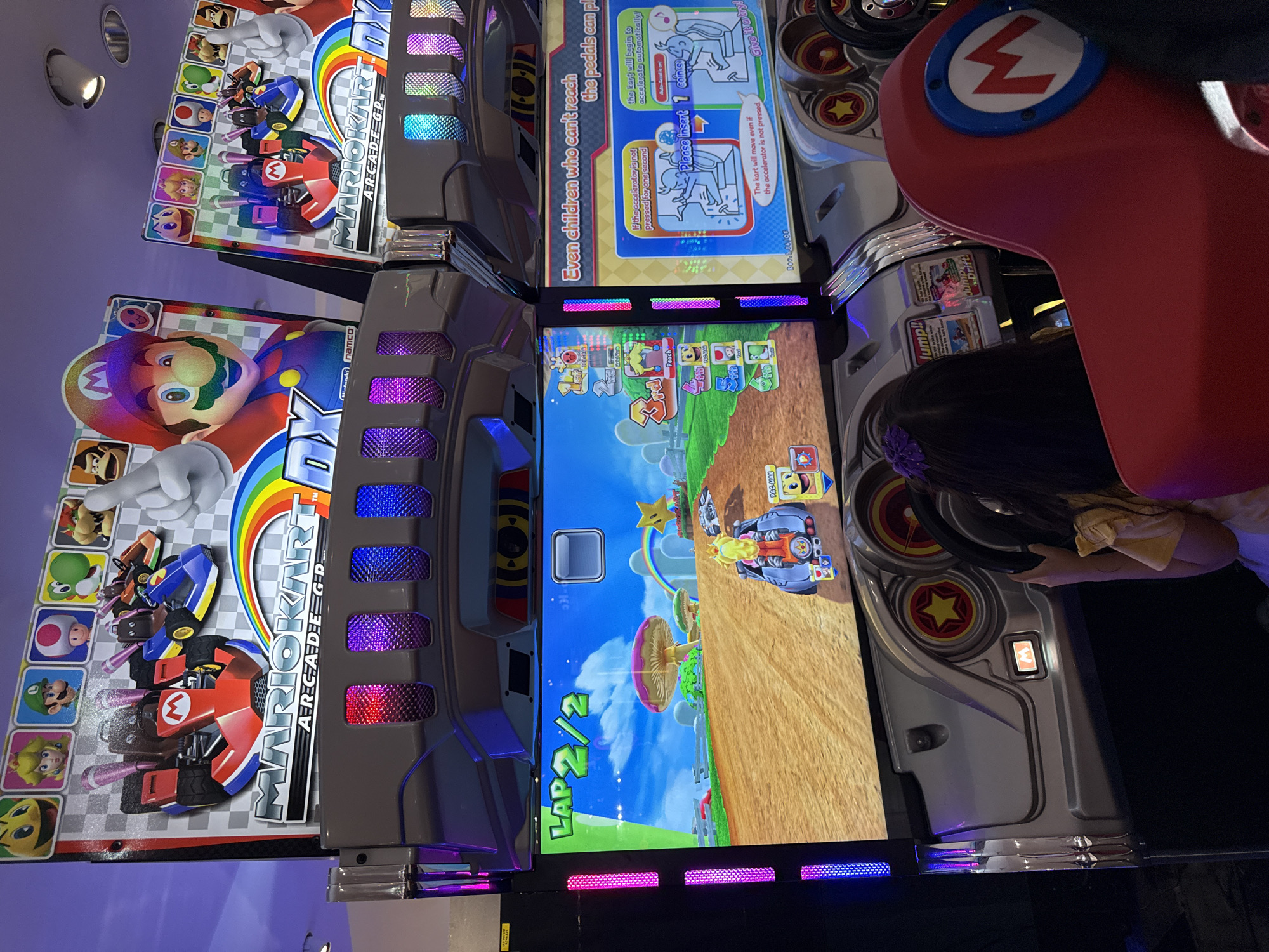 Here is a description of the image:
The image shows a bank of Mario Kart Arcade GP DX games. Three machines are visible in the image. The machines are lit with colorful LED lights and screens displaying the game. One of the machines has a person playing it, with their hands on the steering wheel. The side of each game features an image of Mario Kart characters and logos. The game screens show different levels and characters. Instructions and coin slots are also visible on the machines. The environment is dimly lit, likely inside an arcade. While a specific address is not provided, the picture is described as being taken in Columbus, Ohio.