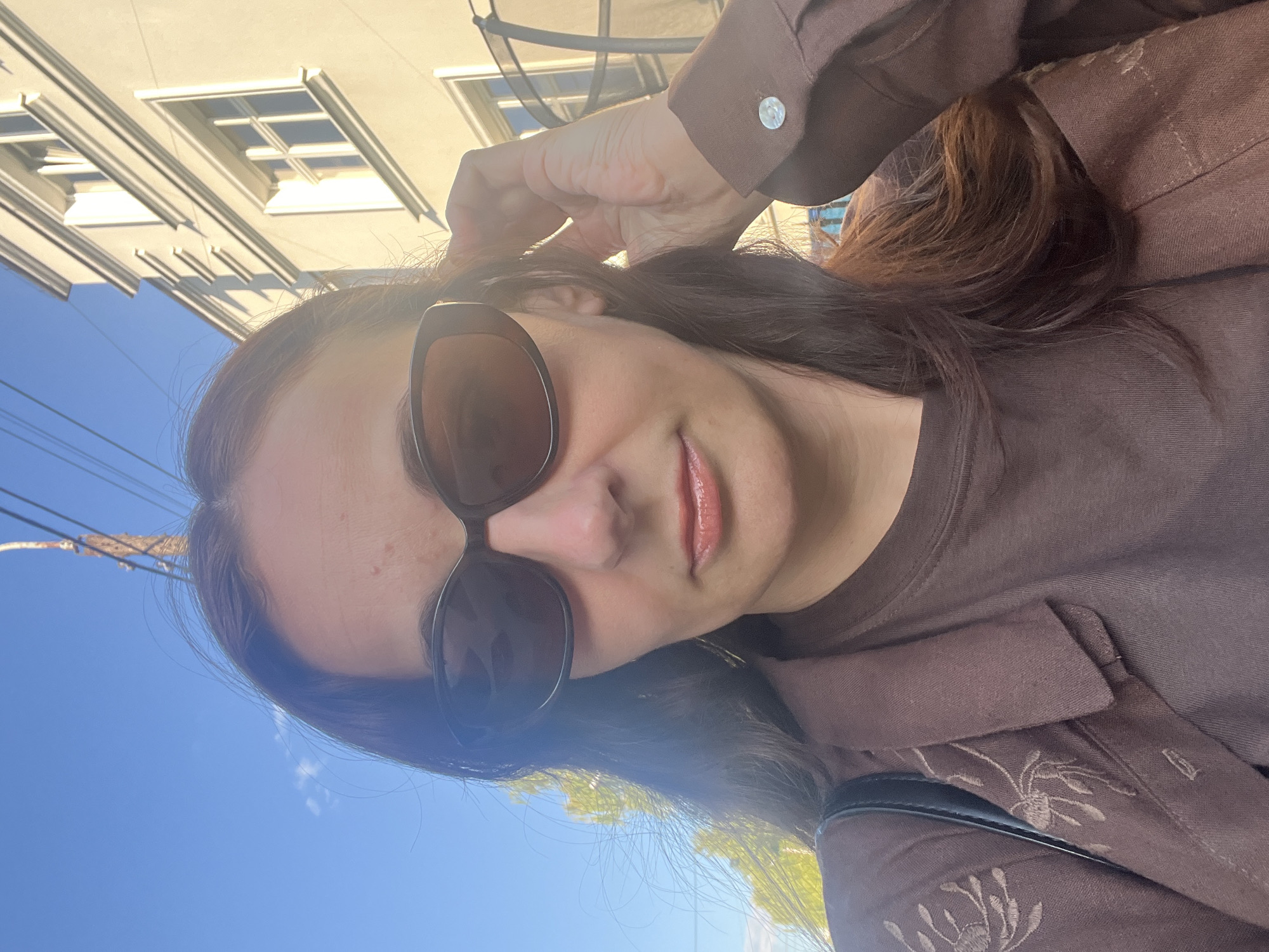 An adult female is depicted in a close-up, outdoor shot under clear blue skies with bright sunlight. She has long, dark brown hair, visible flowing over her shoulders, and wears dark oval-shaped sunglasses that obscure her eyes. Her lips are coated with a glossy, light peach-pink product. She is wearing a dark brown t-shirt beneath a collared, brown jacket or shirt-jacket. The left sleeve of the jacket features subtle embroidered floral patterns in a matching brown thread. Her right hand is raised to the side of her head, with fingers gently touching her hair. A round, light-colored button with a textured surface is visible on the cuff of her right sleeve. A black strap, likely from a bag, is visible over her left shoulder.
The background consists of a clear blue sky with a wispy white cloud in the upper left. A wooden utility pole with multiple black wires runs vertically in the upper left, partially obscured by her head. Green foliage from trees is visible below the wires and in the mid-left. To the right, a light-colored building with architectural moldings and multiple windows is brightly illuminated by the sun, casting distinct diagonal shadows. A dark, possibly metal, structure is partially visible in the far right background behind her hand. Strong lens flare is evident across the sunglasses and the right side of her hair due to the direct sunlight.