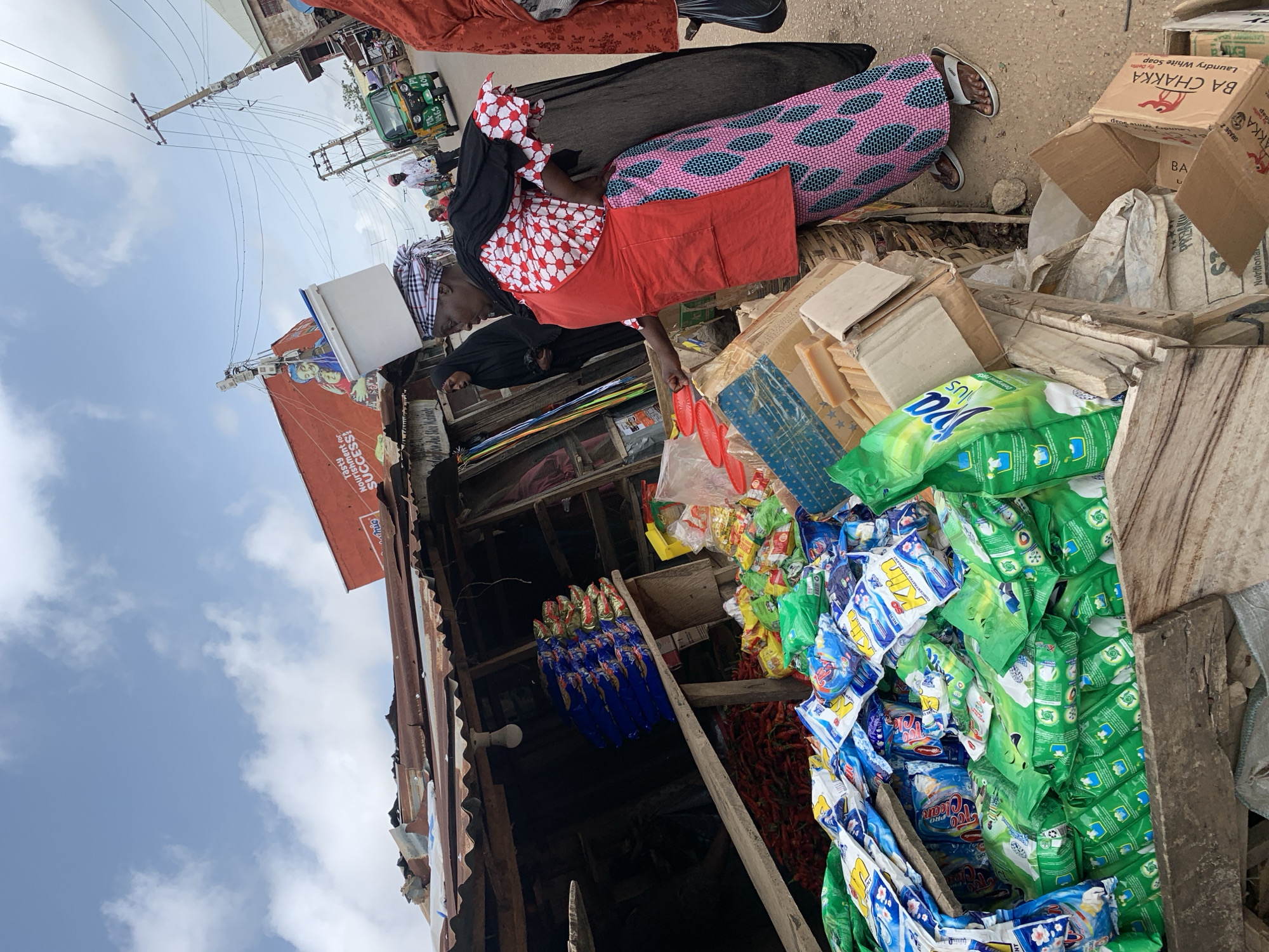 The image depicts a bustling outdoor market scene in Jos, Nigeria, focusing on a woman carrying a white plastic tub on her head. She is dressed in a red apron over a white and red patterned top and a long, patterned pink and blue skirt. Beside her, a stall displays an abundance of colorful detergent packets, primarily green and blue, along with a pile of red chilies and bars of soap. In the background, other market-goers, a green tricycle, and overhead power lines suggest a lively urban environment. The sky above is a mix of blue and scattered clouds, indicating daylight.