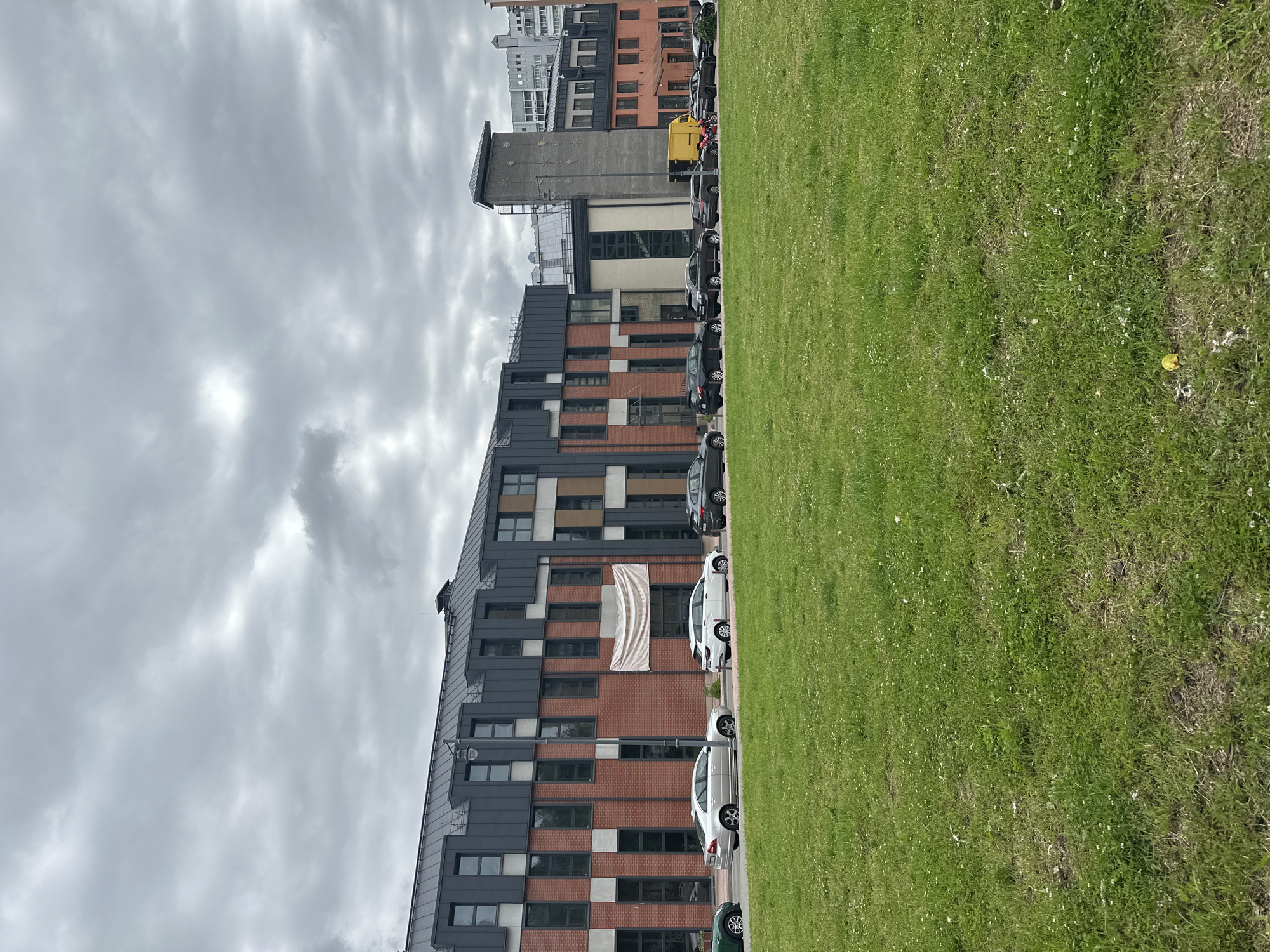 The image shows a row of modern buildings with red brick and dark grey facades, lined with parked cars. A green lawn is in the foreground, and the sky is overcast. A banner hangs from one of the buildings. The scene appears to be a street or parking area in front of an urban development. This location is identified as Minsk, Belarus.